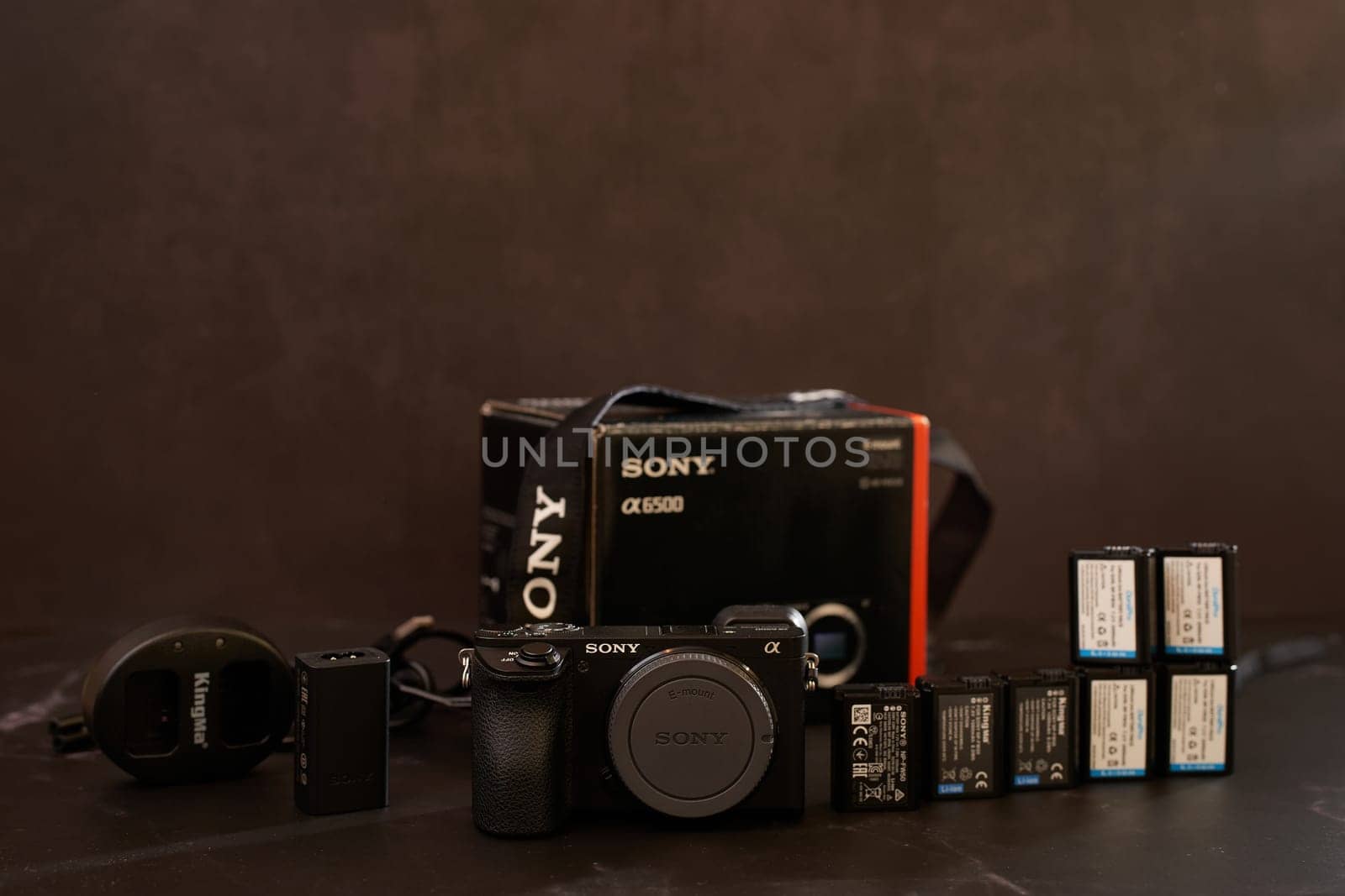 Sony A6500 mirrorless camera with charger, batteries and strap stands on a black table. High quality photo