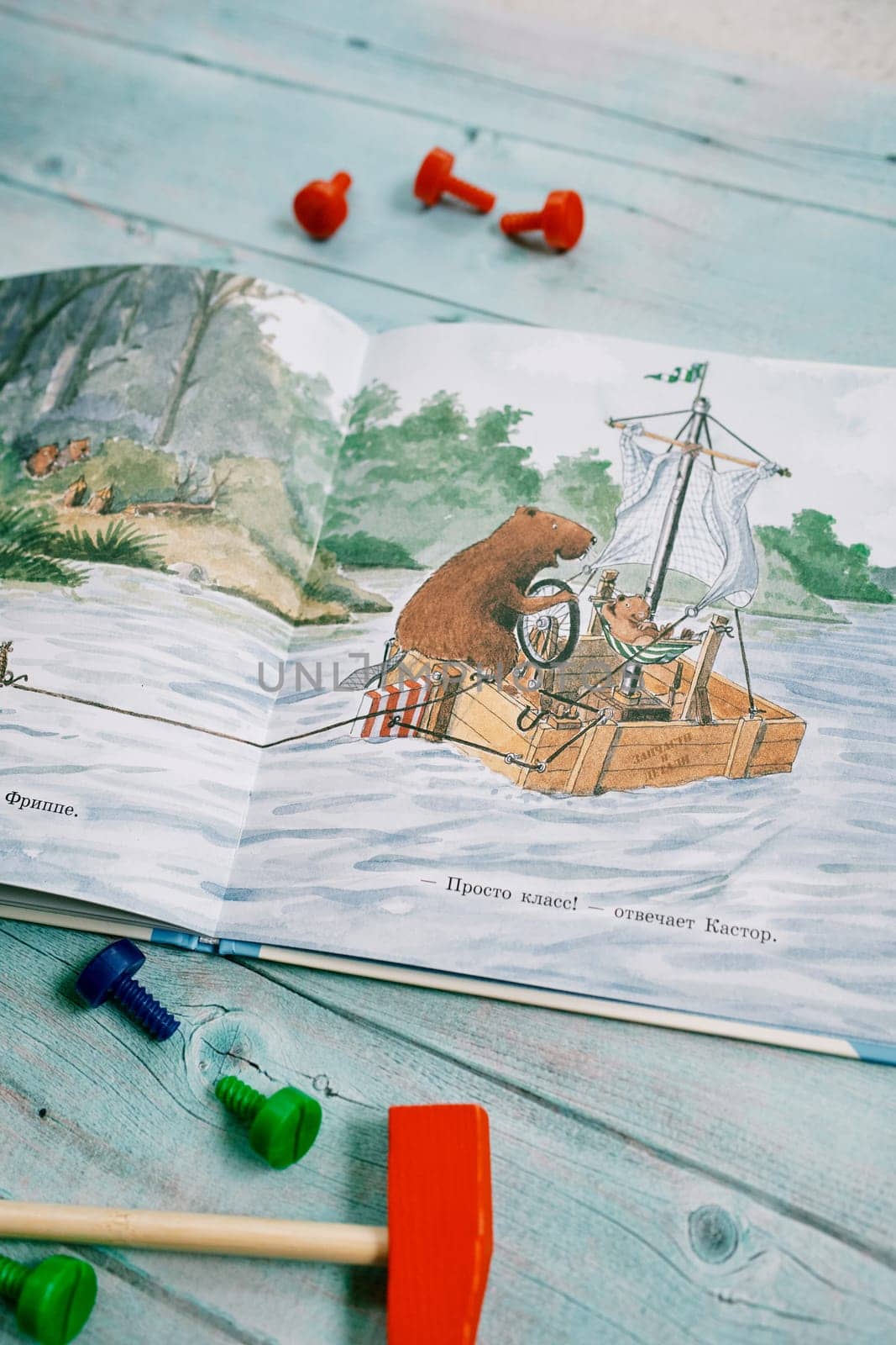 Open book of fairy tales with drawn beavers sailing in a boat lies on a wooden table next to a toy hammer and bolts. High quality photo