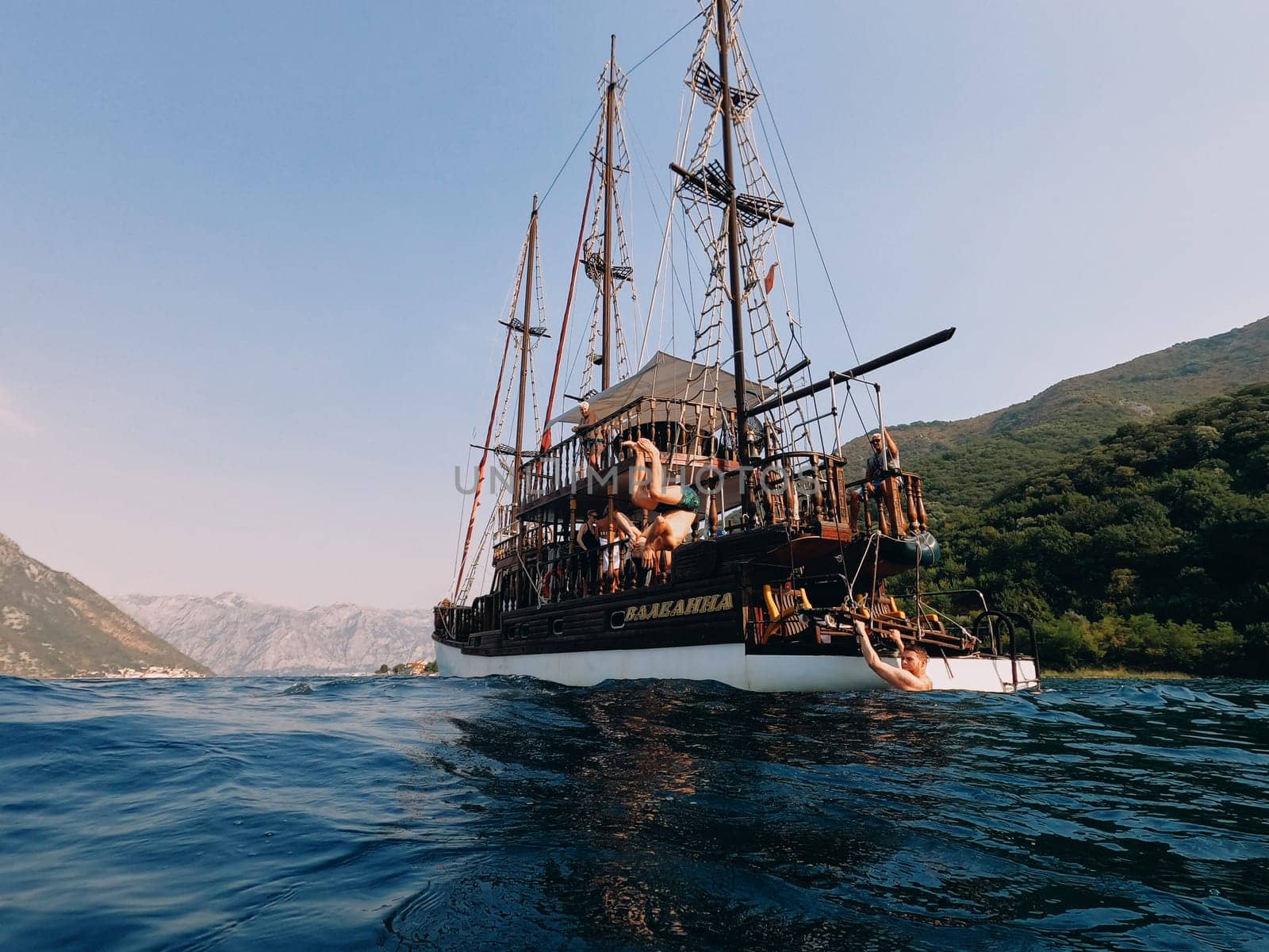 Guys jumping into the sea from the deck of a wooden sailing yacht standing near the mountainous coast. High quality photo