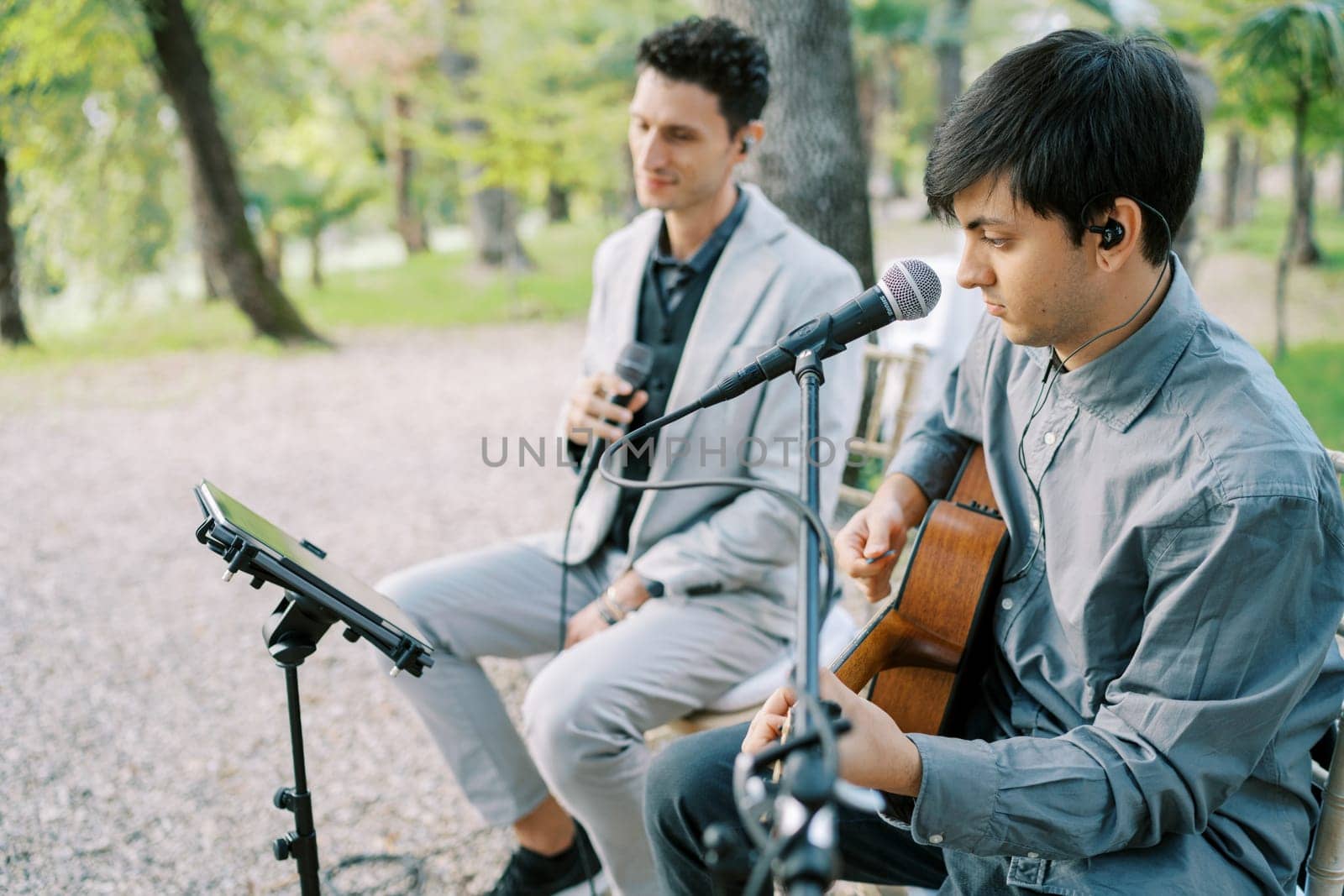 Musician plays an acoustic guitar while sitting on a chair in a park next to a singer. High quality photo