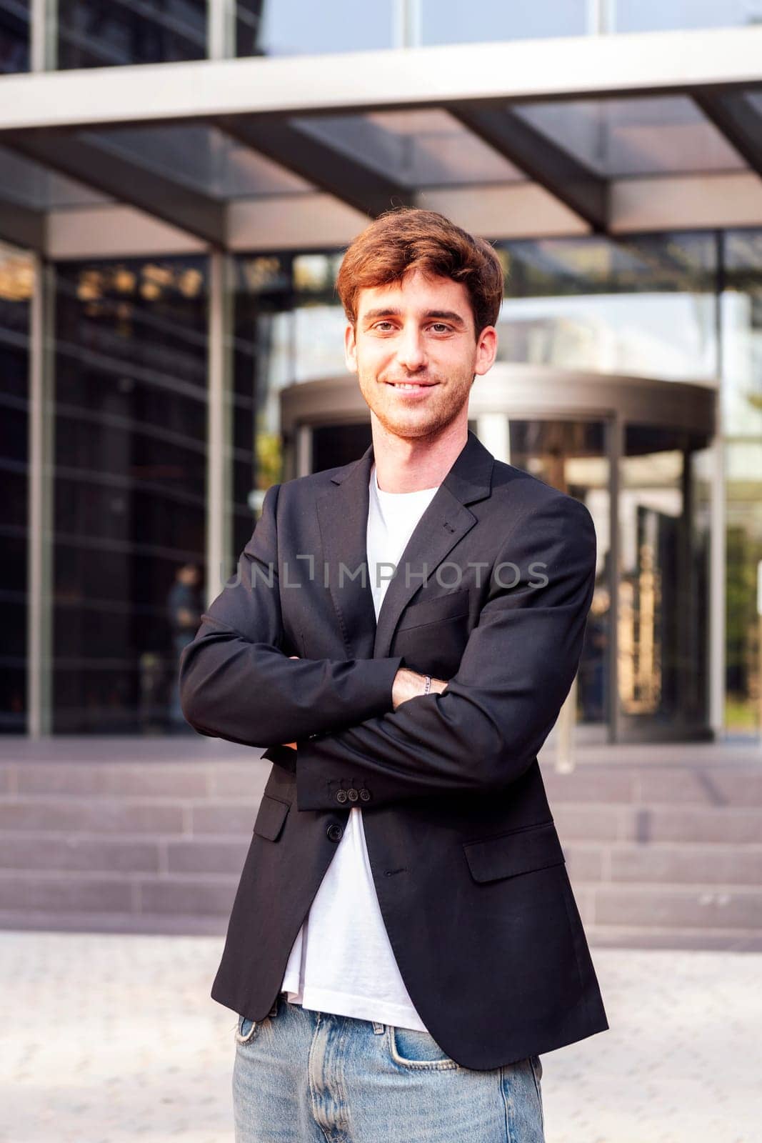 young adult man in casual suit standing looking at camera with arms crossed in front of an office building, concept of entrepreneur and business, copyspace for text