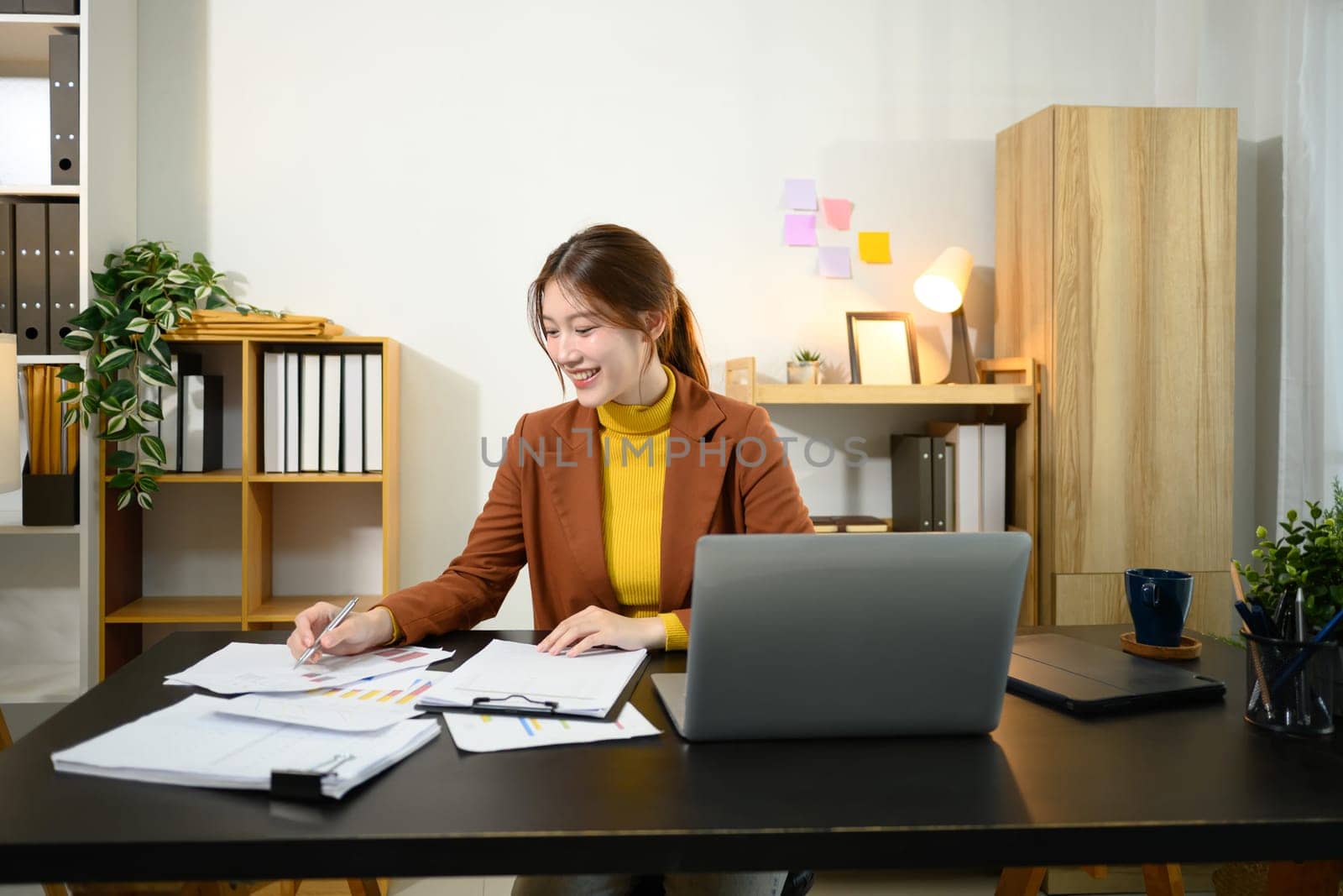 Smiling businesswoman sitting at desk with laptop, reviewing graphs and statistics.