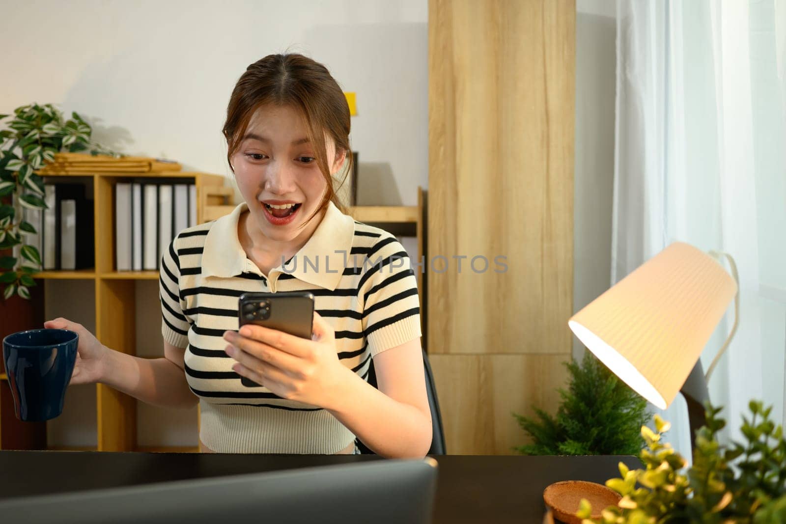 Excited young woman holding a coffee mug and reacting to good news on her smartphone.