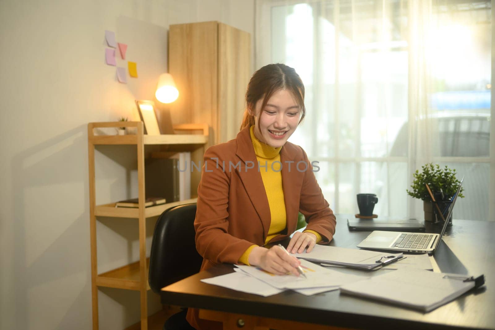 Smiling businesswoman sitting at desk with laptop, reviewing graphs and statistics.