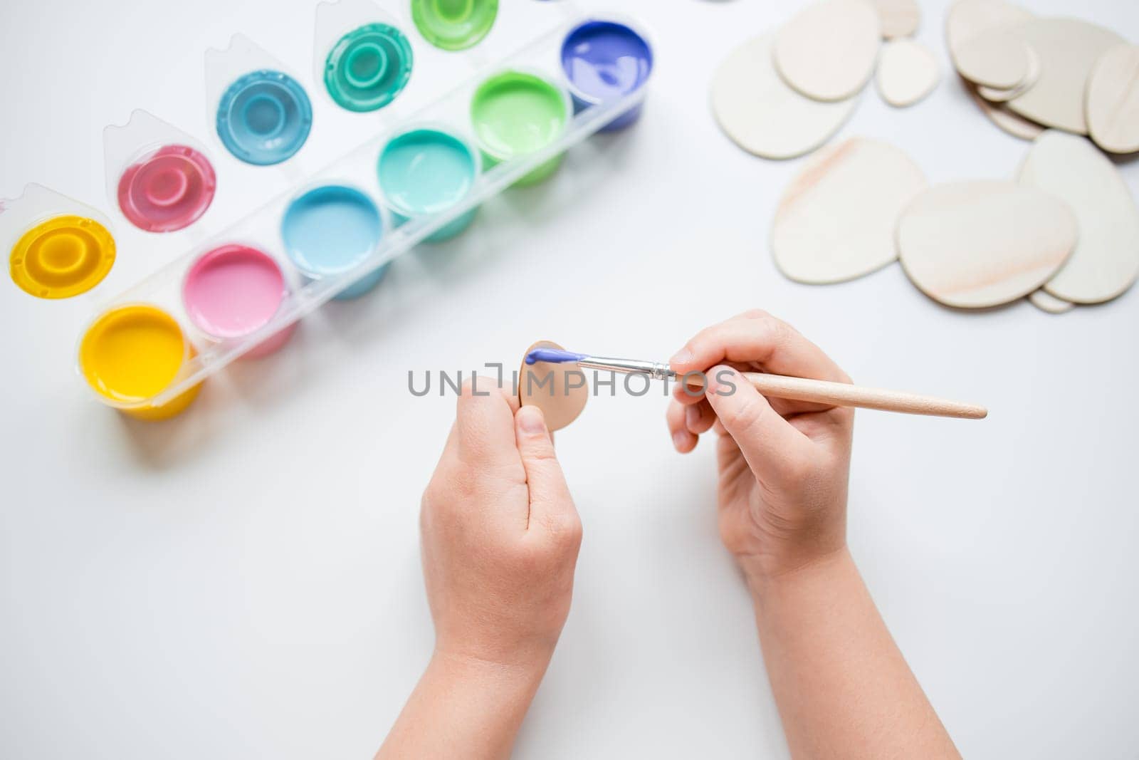 Child painting Esater wooden egg shapes with colorful palette on white table.
