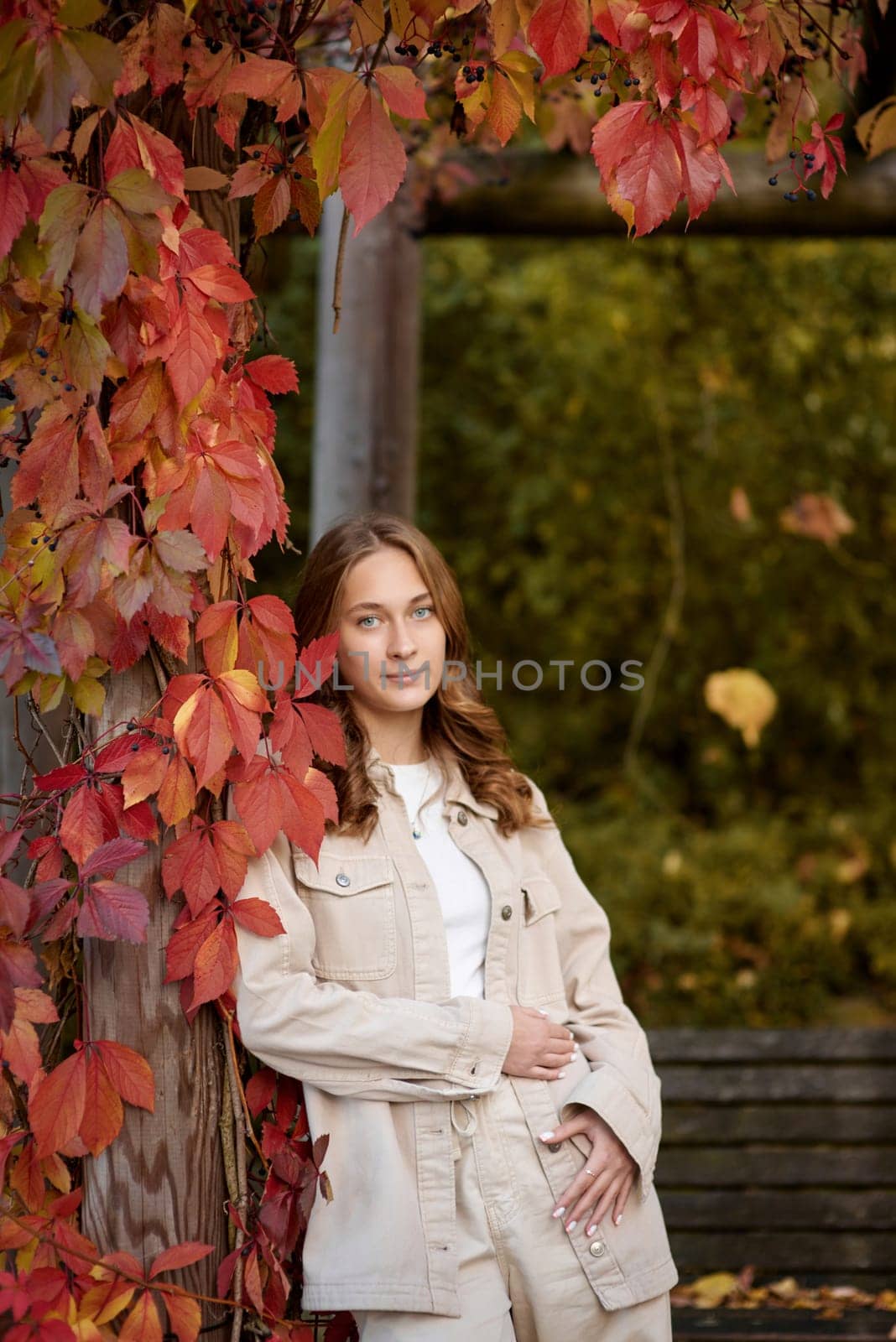 A radiant young woman in a casual beige ensemble leans gracefully against a rustic wooden pergola adorned with vibrant red autumn leaves. The warm, golden tones of fall foliage create a natural backdrop, highlighting her joyful smile and the inviting atmosphere of the garden setting. This image captures the essence of autumn lifestyle, combining modern casual fashion with the organic textures of seasonal nature. Perfect for promoting outdoor living, fashion blogs, travel content, or autumn-themed marketing campaigns, the scene evokes feelings of relaxation, connection with nature, and the comforting charm of the fall season. A young woman with wavy light brown hair and a cheerful smile is leaning against a wooden pillar wrapped in red autumn leaves. She is dressed in a beige casual outfit, including a stylish denim jacket and matching pants, with a white top underneath. The background features a scenic garden pergola with wooden beams covered in lush green and red foliage, creating a natural and cozy atmosphere. The warm lighting and soft depth of field enhance the autumn aesthetic, making this image perfect for seasonal fashion, lifestyle blogs, outdoor portraiture, and autumn-themed marketing campaigns.