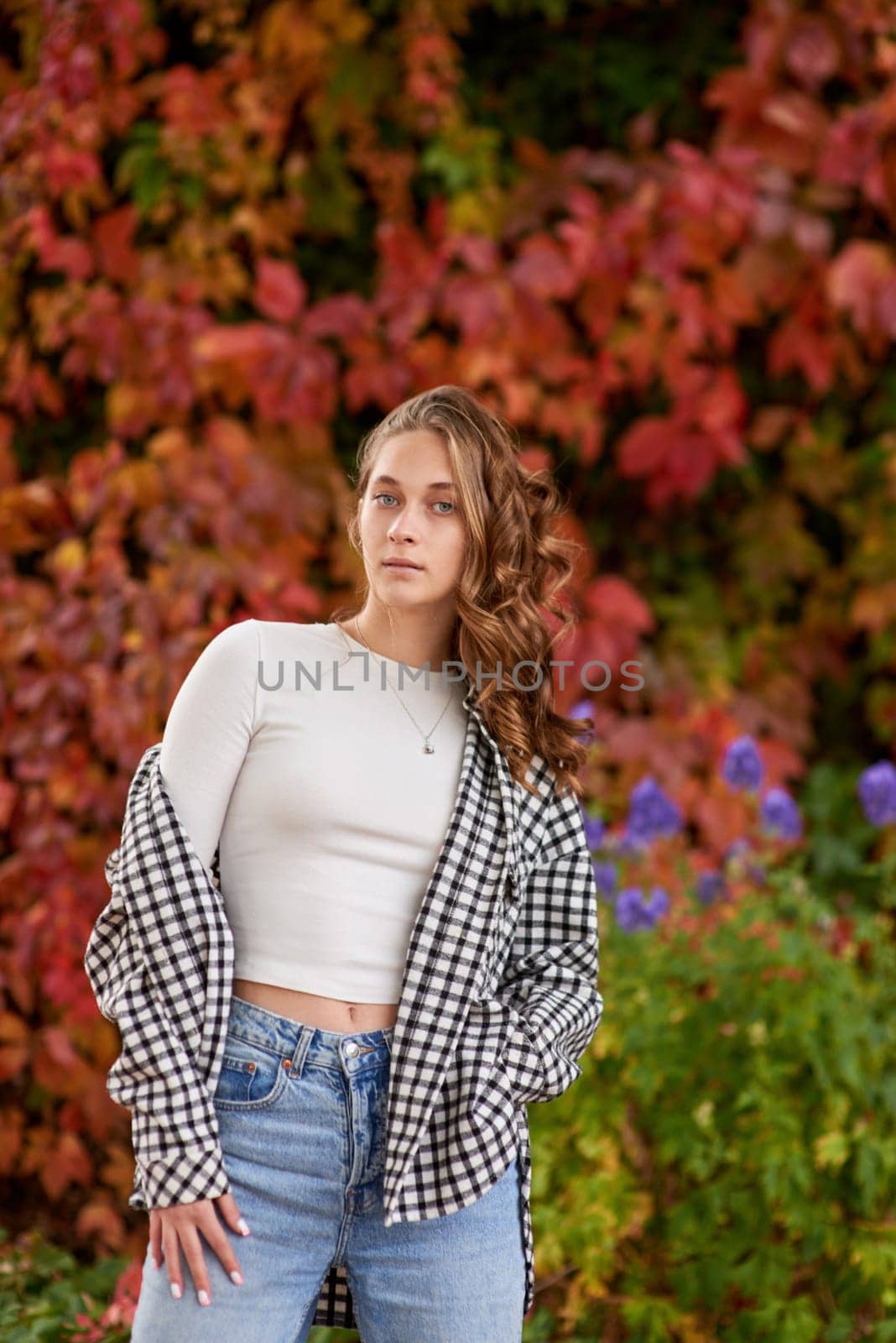 A young woman in a casual autumn outfit poses confidently against a vibrant red ivy wall, creating a stylish fashion portrait in warm seasonal tones. The cozy fall atmosphere and urban outdoor setting highlight trendy seasonal style, making this image perfect for fashion blogs, lifestyle content, seasonal marketing campaigns, or travel-related projects. The rich red tones of the ivy wall, combined with the natural light and relaxed vibe, evoke a sense of warmth, elegance, and modern urban charm.