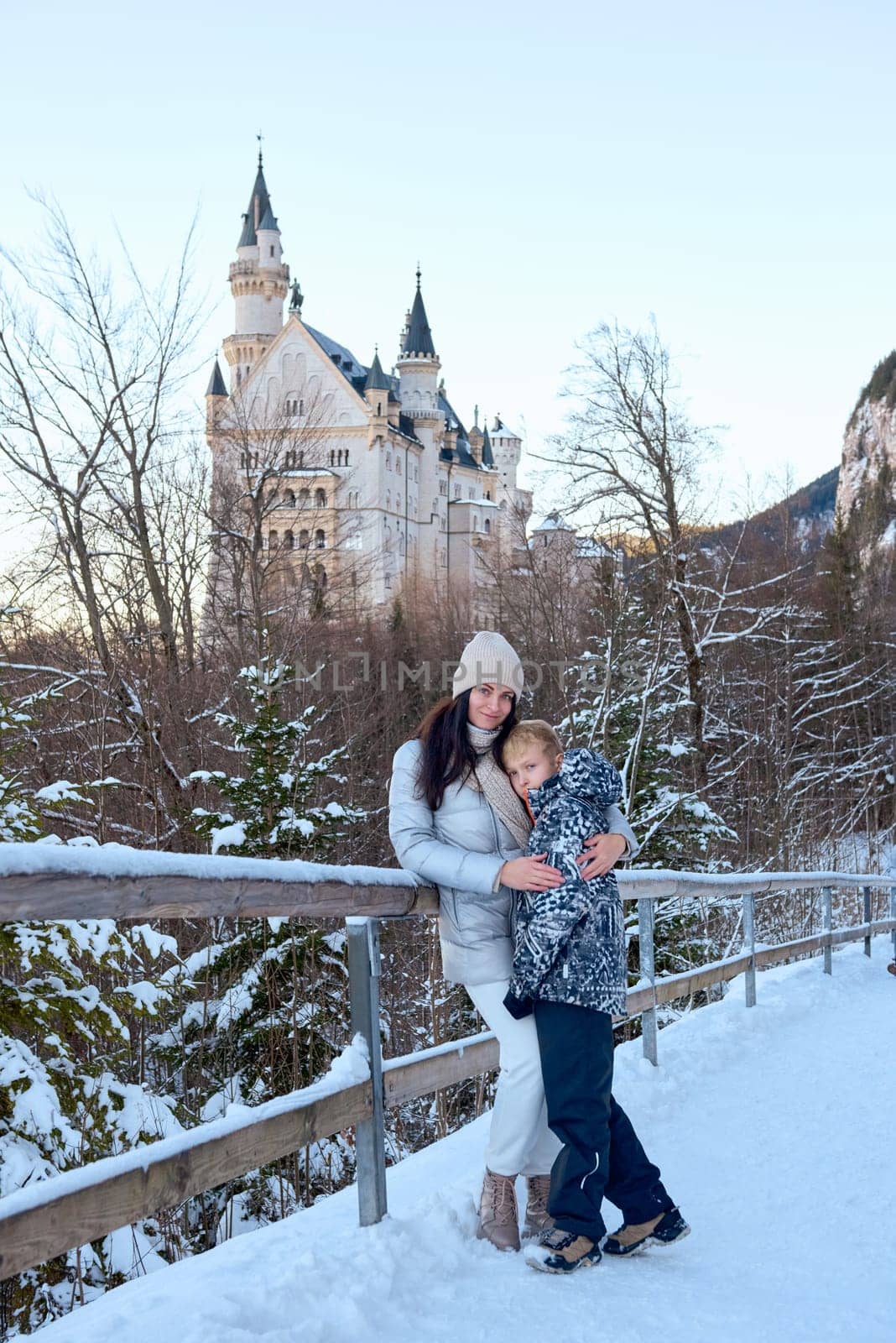This photograph captures a beautiful woman and a child standing on a bridge in the snow, with the iconic Neuschwanstein Castle prominently featured in the background. The scene exudes a serene and picturesque winter atmosphere, with the majestic castle and the snowy landscape creating a stunning backdrop. The woman and child are dressed in warm winter clothing, adding to the cozy and inviting feel of the image. The photograph is perfect for themes related to family, winter travel, nature, and scenic landscapes, offering a detailed and evocative representation of a winter wonderland. Beautiful woman and child standing on a bridge in the snow with Neuschwanstein Castle in the background, capturing a serene winter scene with a majestic castle and snowy landscape.