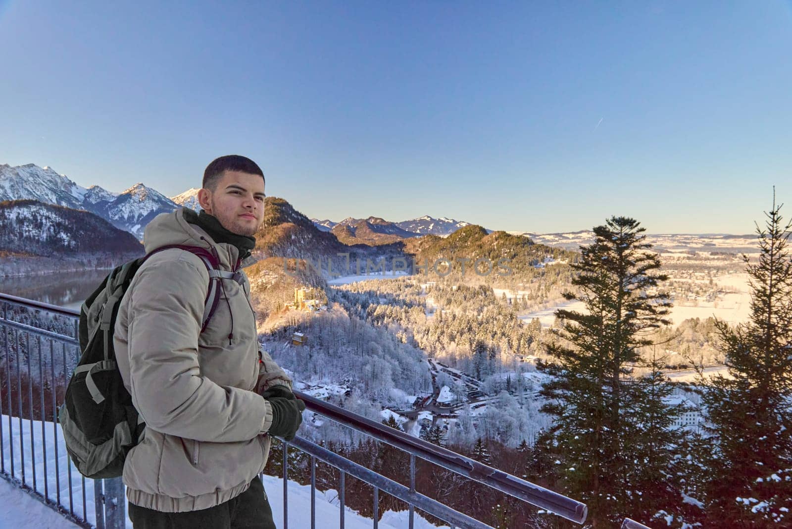 A striking image of a man standing confidently on a balcony, gazing out at the majestic icy mountains and a lush forest in the background. The photo is taken in portrait mode, capturing the breathtaking winter landscape with towering snow-capped mountains and the serene forest below. The man, dressed warmly for the cold weather, stands in front of the dramatic mountain range, adding a human element to the stunning natural scene. The icy peaks reflect the light, while the surrounding forest enhances the depth of the shot. This captivating image evokes a sense of adventure and awe, perfect for winter tourism, outdoor exploration, and scenic landscape photography Man standing on a balcony with icy mountains and a forest in the background, breathtaking scenic view, portrait mode, winter landscape photography