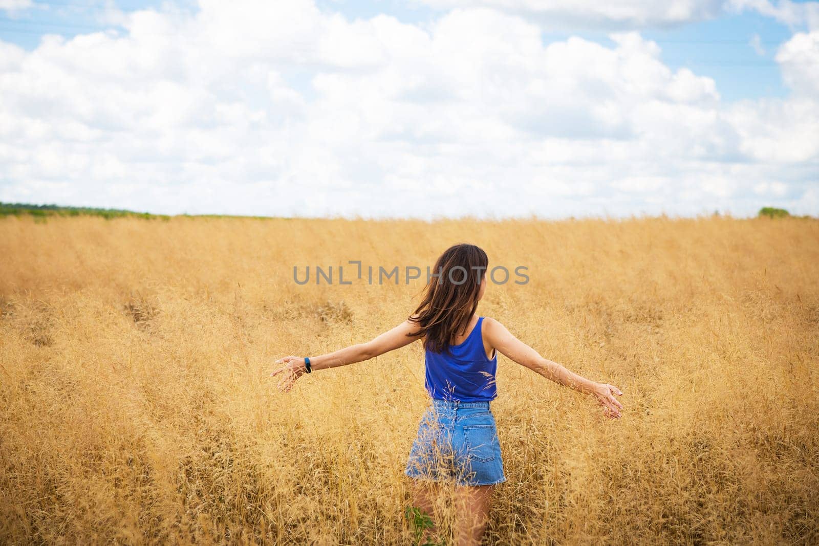 Portrait of a young beautiful brunette woman in a blue T-shirt and denim shorts stands in the middle of the field, exposing her face to the sun on the background of a summer day outdoors