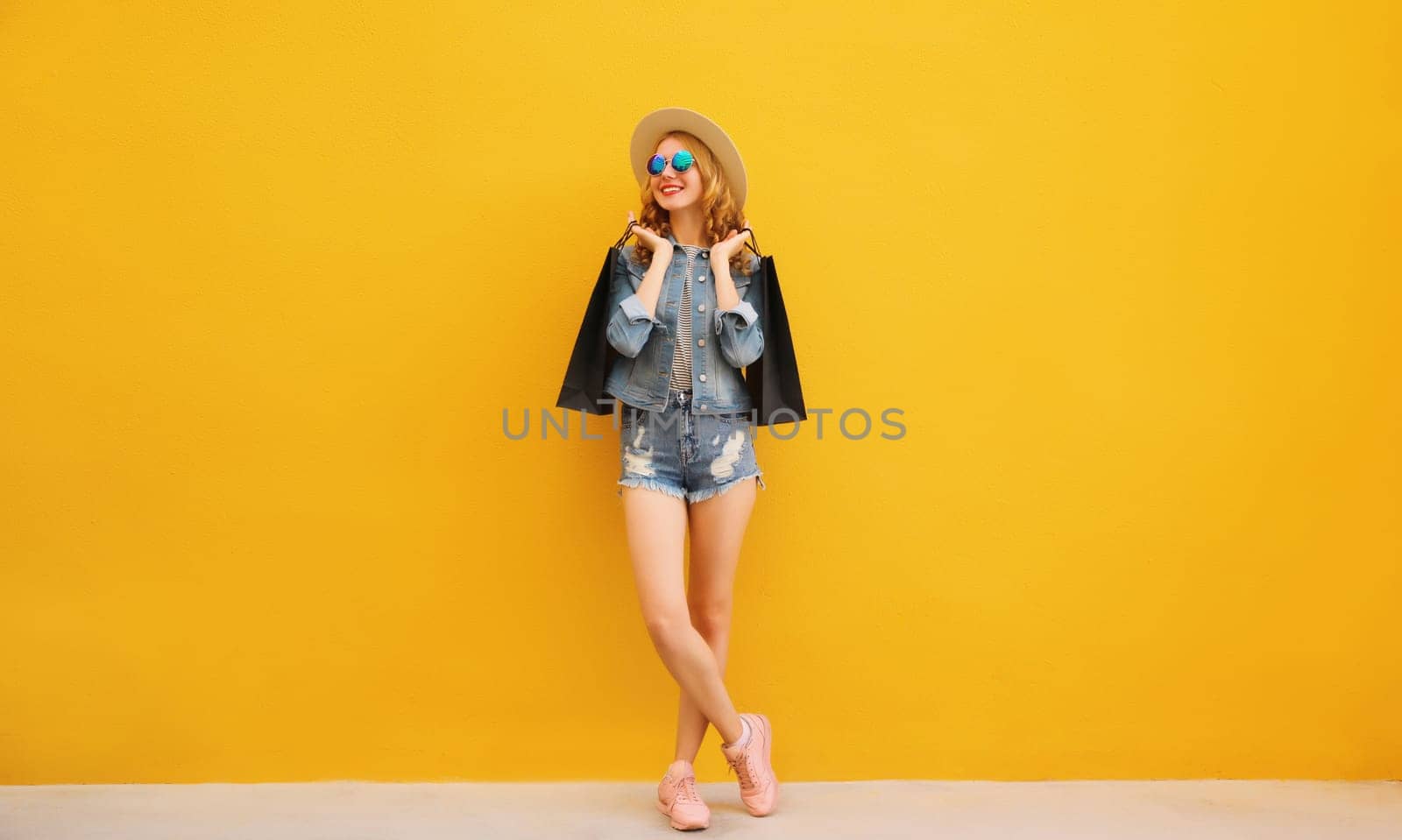Stylish beautiful happy smiling young woman posing with black shopping bags walking looking away wearing denim clothing, summer straw hat on yellow studio background