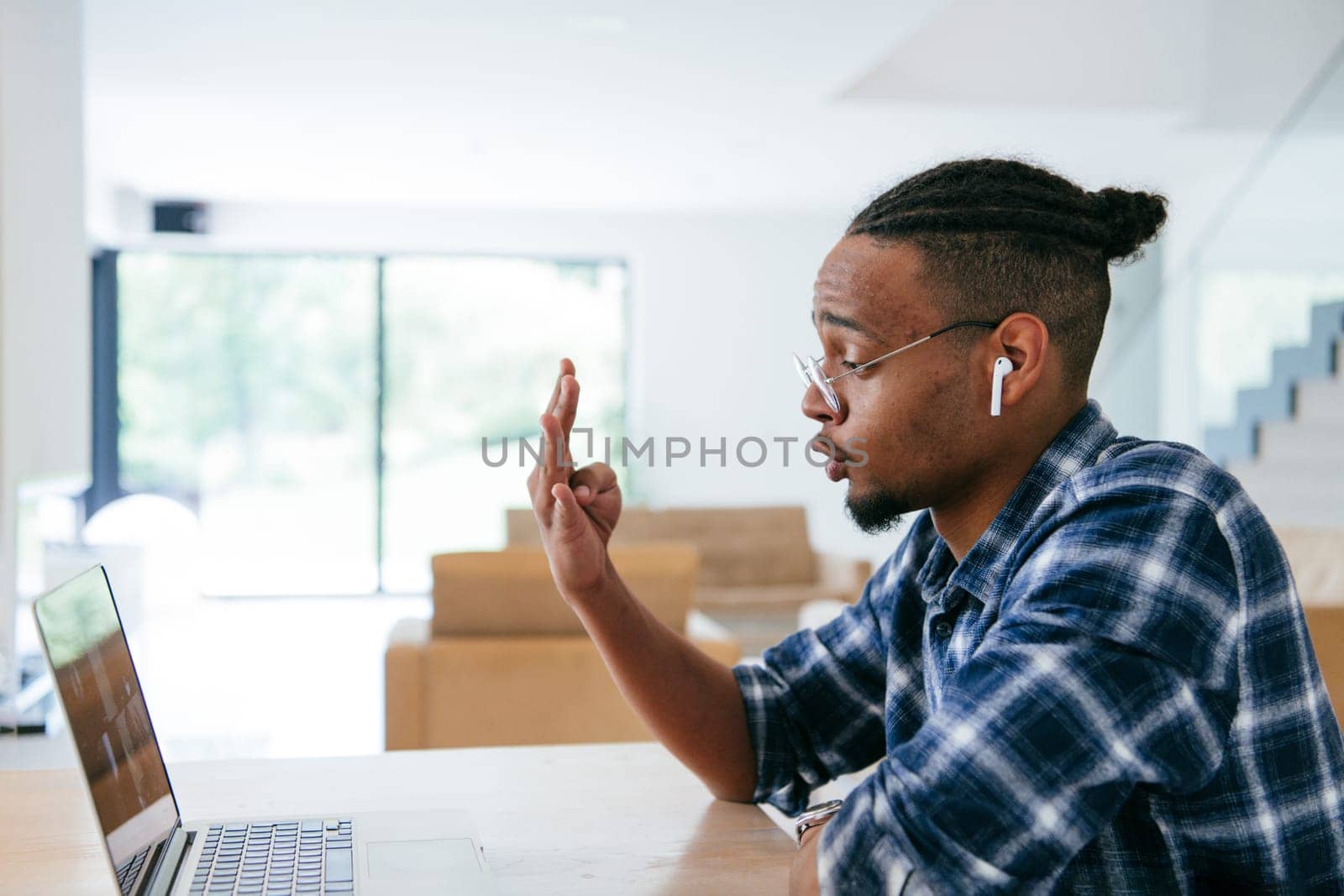 African American influencer in glasses sitting at a table in a modern living room, using a laptop for business video chat, conversation with friends and entertainment