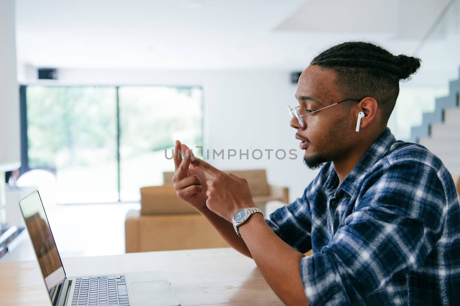 African American influencer in glasses sitting at a table in a modern living room, using a laptop for business video chat, conversation with friends and entertainment