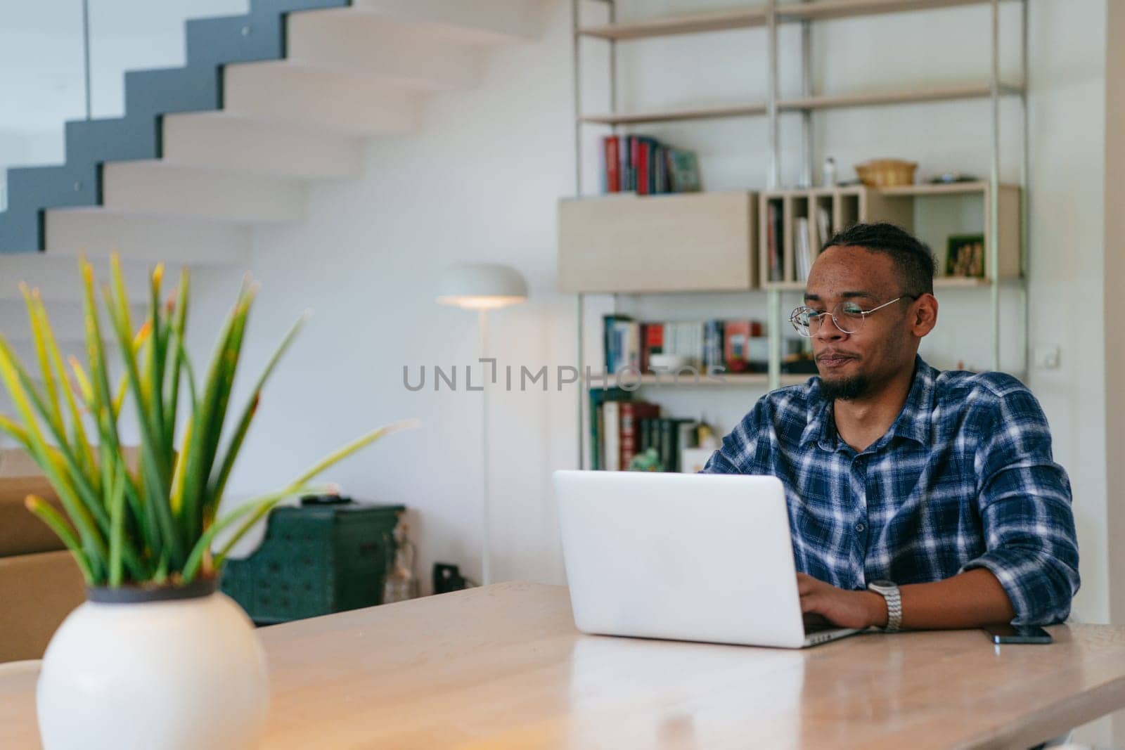 African American influencer in glasses sitting at a table in a modern living room, using a laptop for business video chat, conversation with friends and entertainment