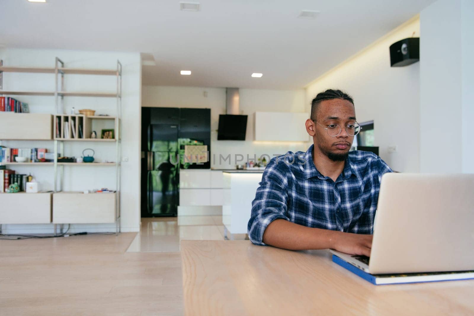 African American influencer in glasses sitting at a table in a modern living room, using a laptop for business video chat, conversation with friends and entertainment