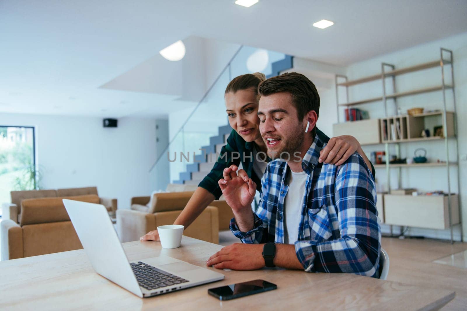 A young married couple is talking to parents, family and friends on a video call via a laptop while sitting in the living room of their modern house
