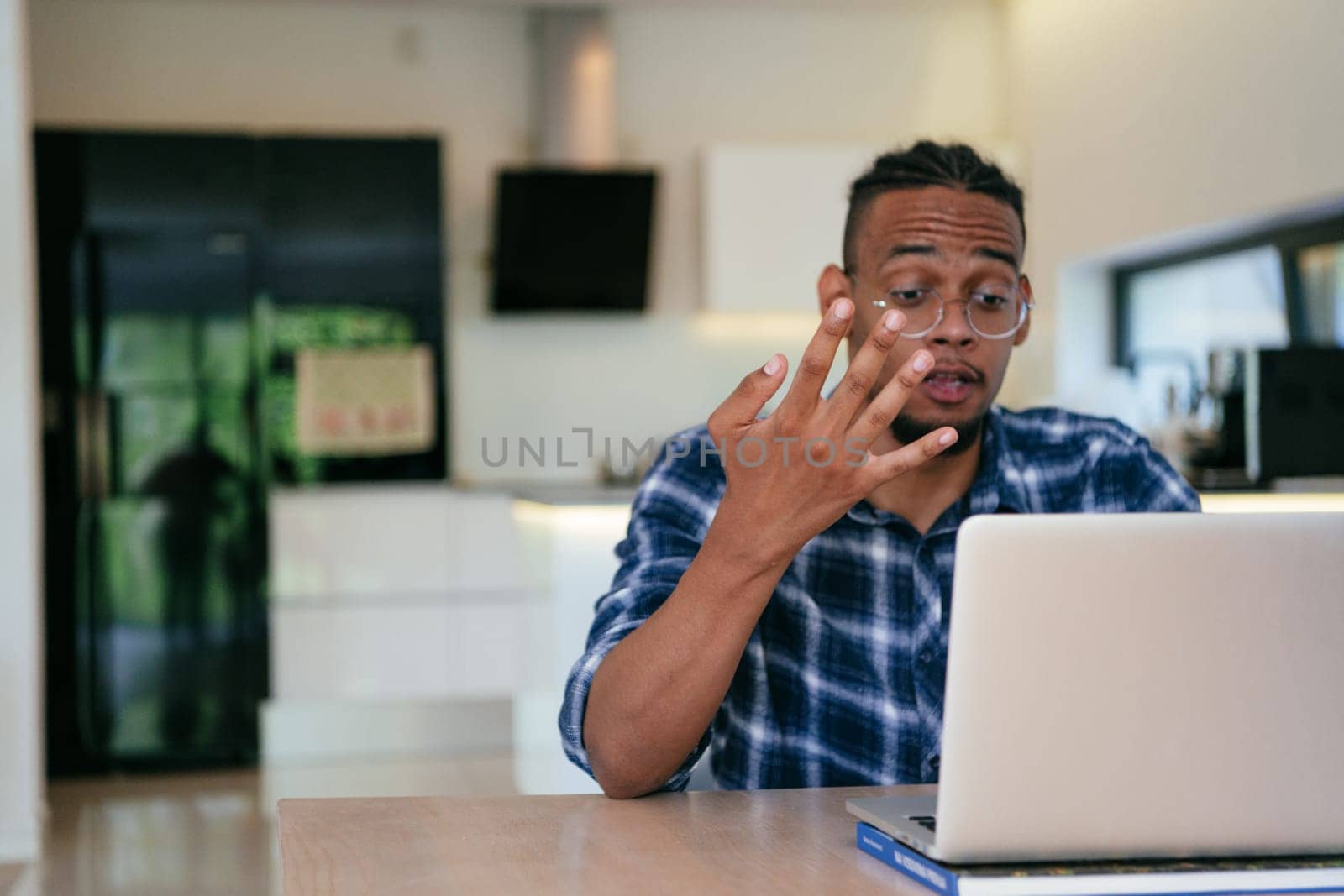 African American influencer in glasses sitting at a table in a modern living room, using a laptop for business video chat, conversation with friends and entertainment