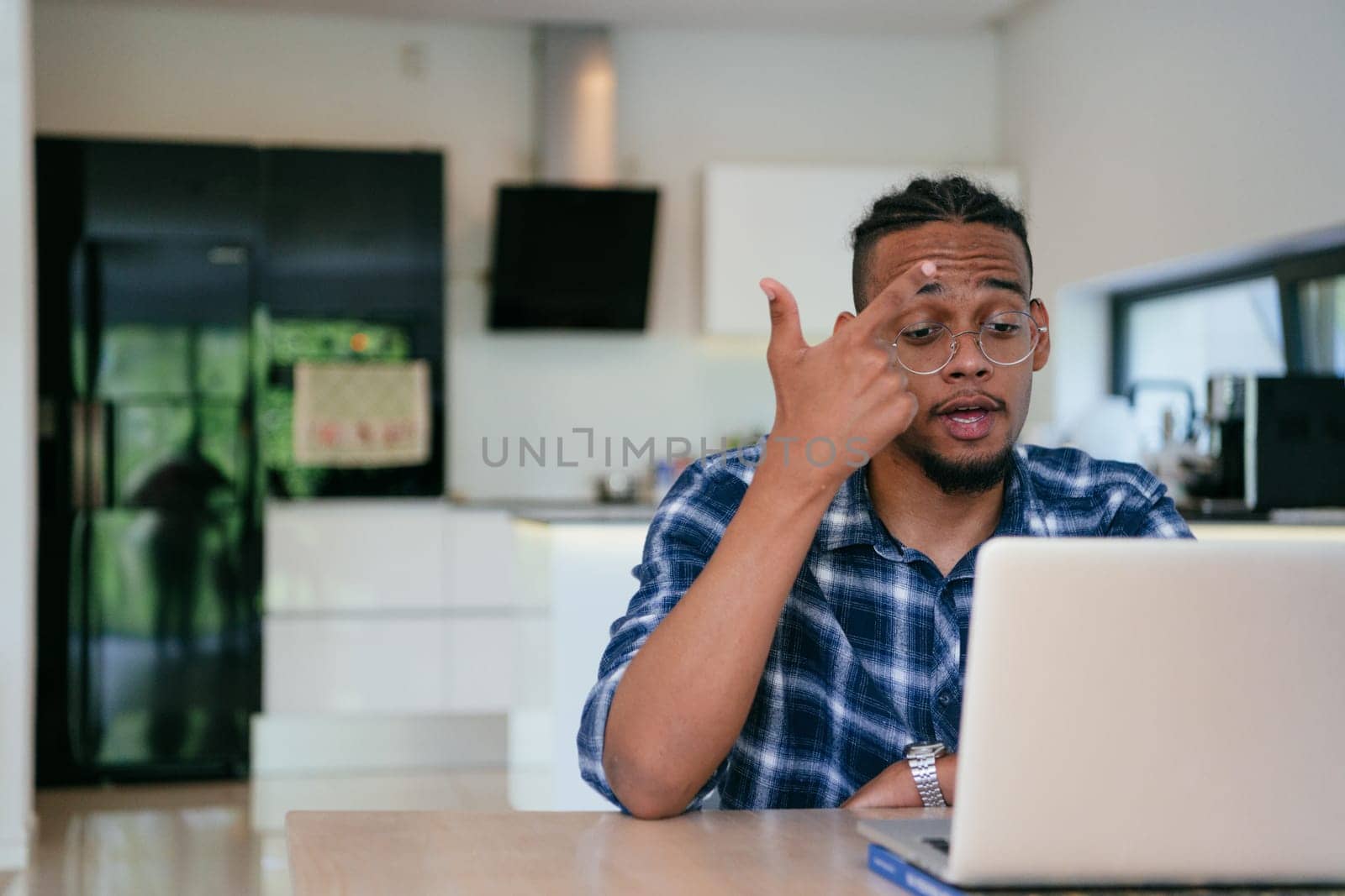 African American influencer in glasses sitting at a table in a modern living room, using a laptop for business video chat, conversation with friends and entertainment