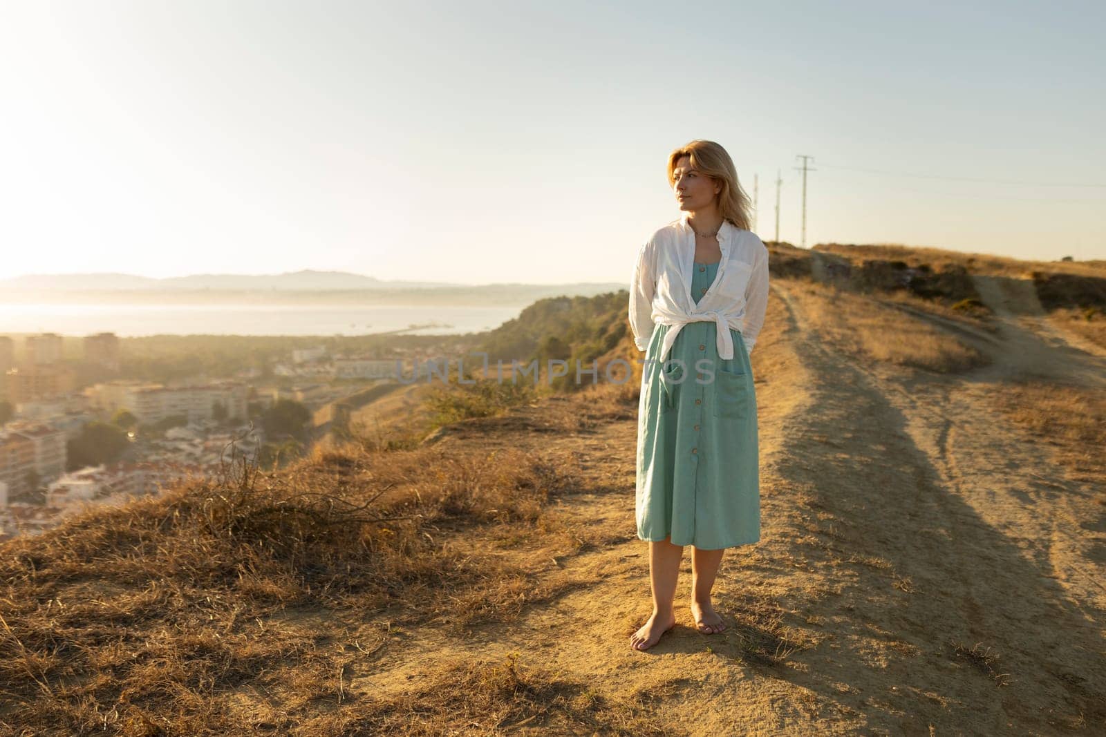 Woman standing barefoot on a hilltop, enjoying a breathtaking sunset view over a coastal city and the sea