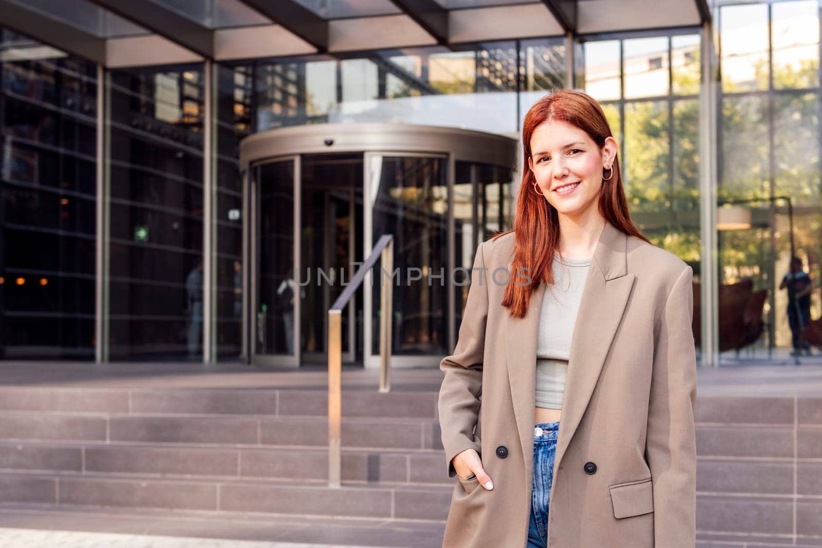 smiling young adult woman in casual suit standing looking at camera in front of an office building, concept of entrepreneur and business