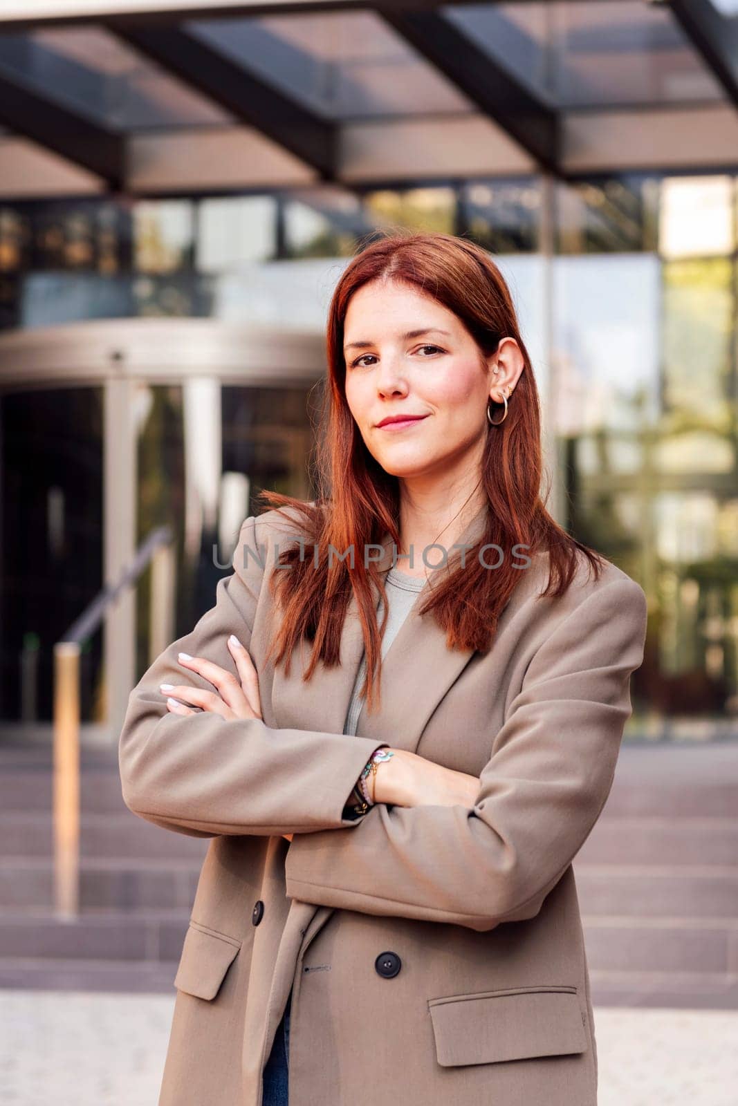 smiling young adult woman in casual suit standing looking at camera with arms crossed in front of an office building, concept of entrepreneur and business, copyspace for text