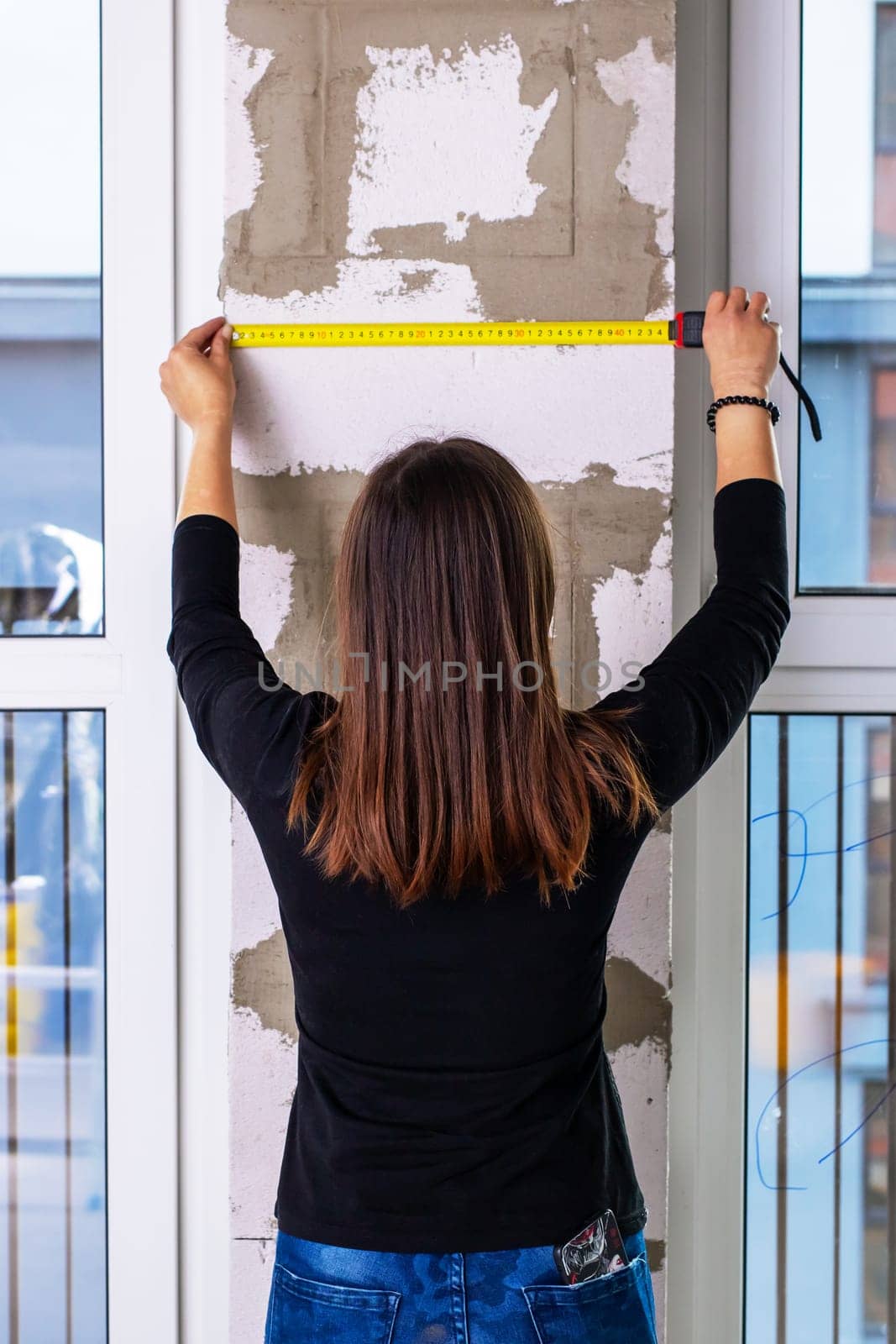 A woman diligently measures a walls dimensions using a tape measure, ensuring accuracy for her upcoming project