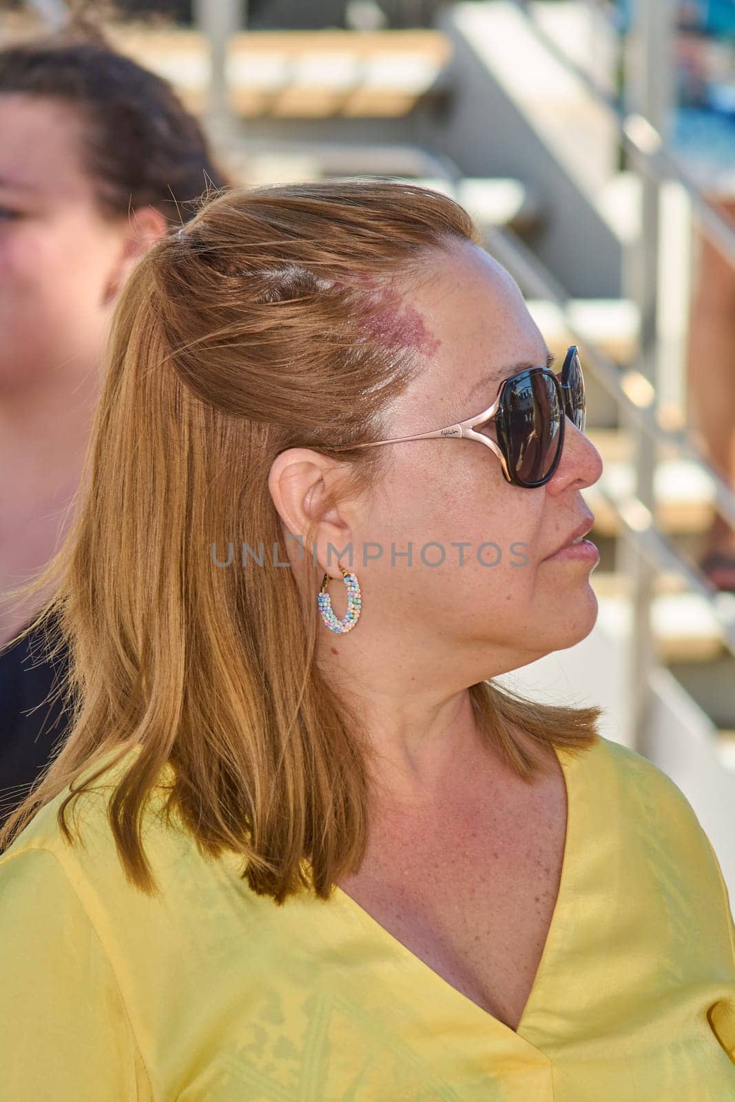 Portrait of a blonde woman with eyeglasses and colorful earrings, dressed in bright yellow. Outdoor setting with blurred natural elements.