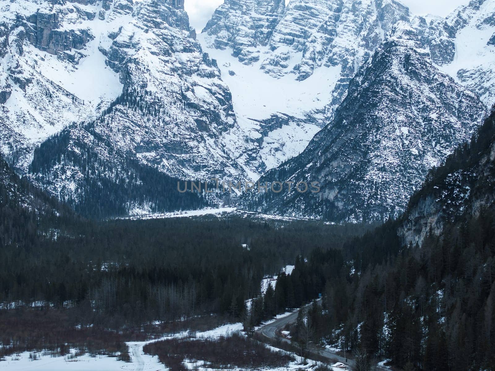 A frozen lake in the Alps is surrounded by dense evergreen forests and towering snow covered peaks. A winding road leads towards the serene landscape.