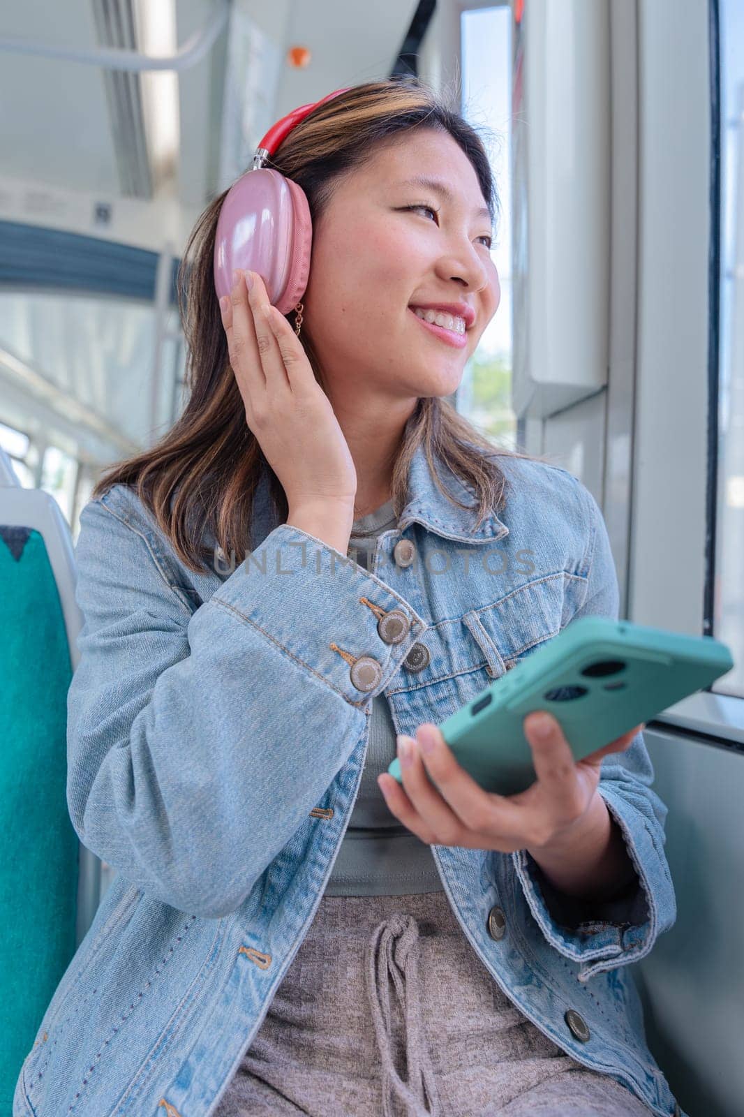 Young Chinese woman with pink headphones enjoying music on tram, eyes closed in relaxation. She holds her smartphone while leaning back in her seat, urban vibe and peaceful moment.Vertical