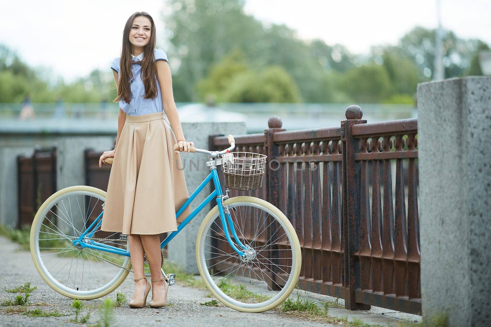 Stylish young woman with long hair smiling, posing with a vintage blue bicycle on a city bridge, wearing an elegant beige skirt and heels