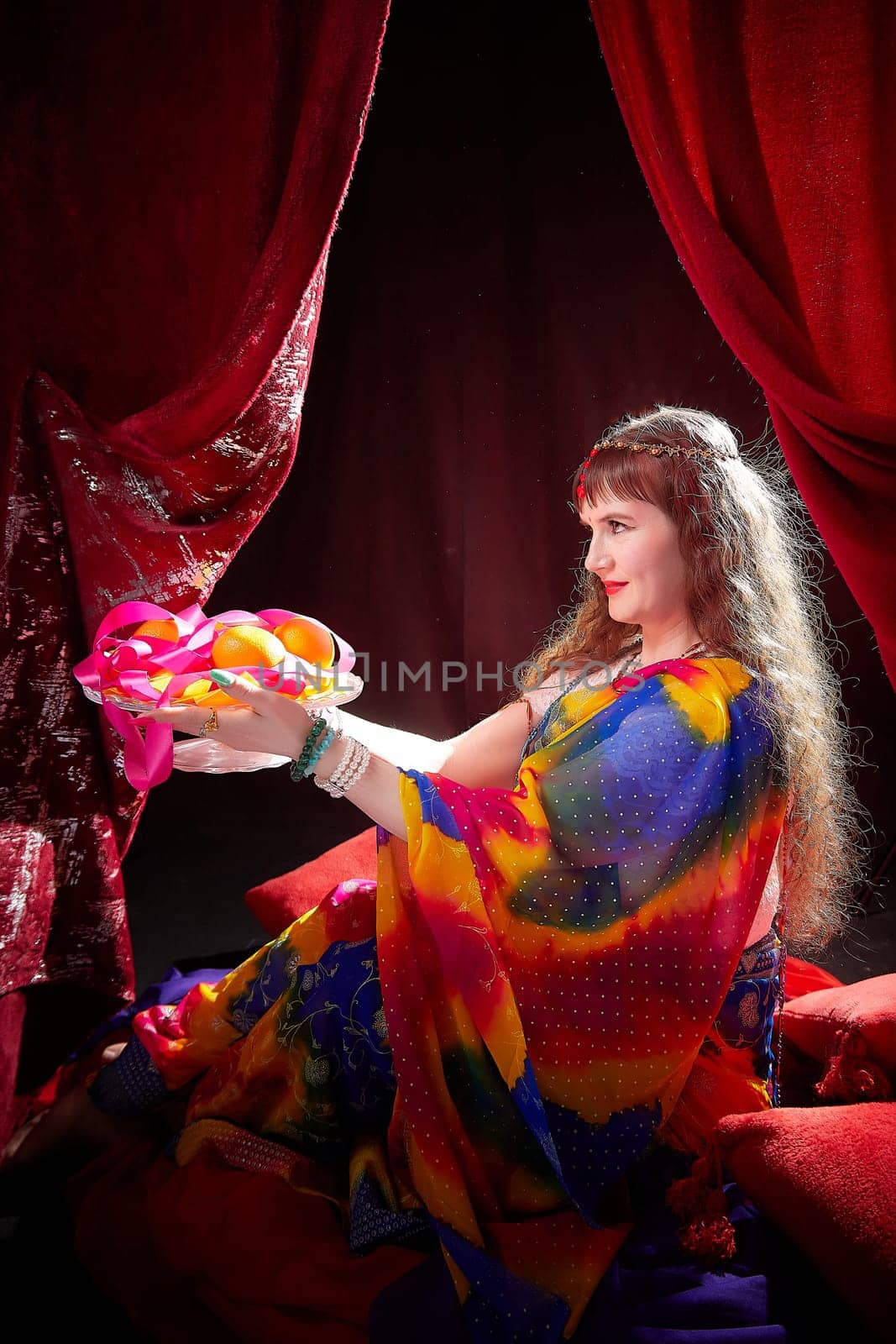 A mature woman dressed in a colorful sari poses gracefully while holding a plate of fruits in a rich Indian backdrop.