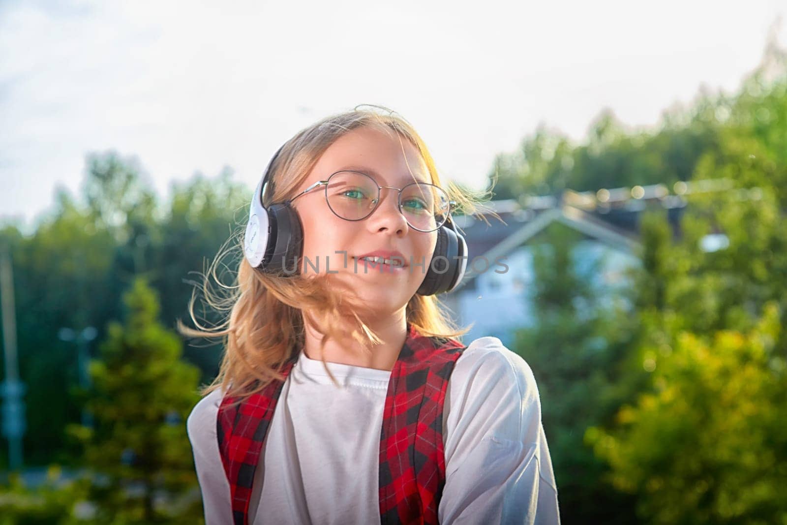 A young girl listens to music while standing outside, wearing headphones and smiling in a vibrant, sunny environment.