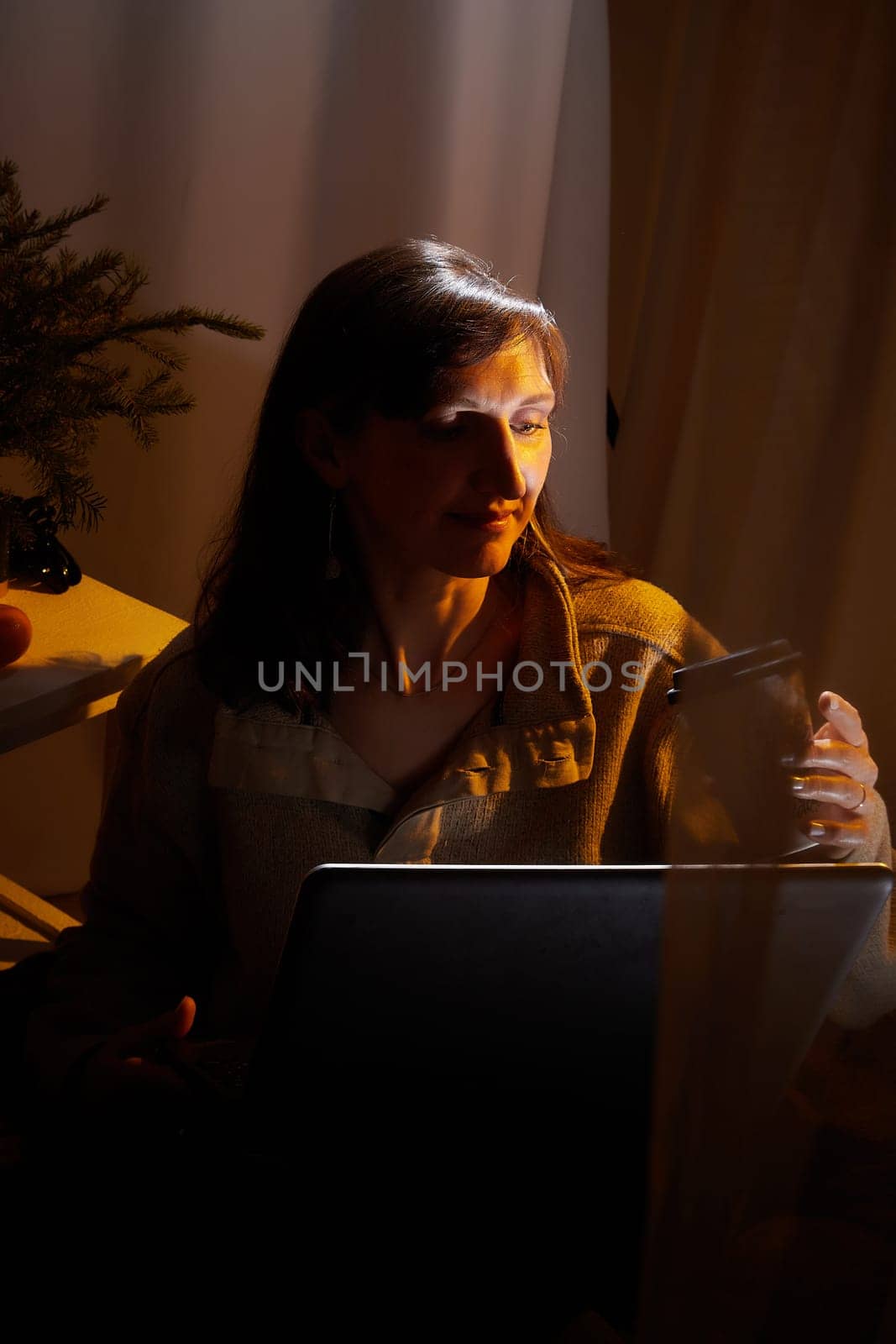 A middle-aged woman relaxes in her cozy apartment during the evening, enjoying a warm drink while using her laptop.