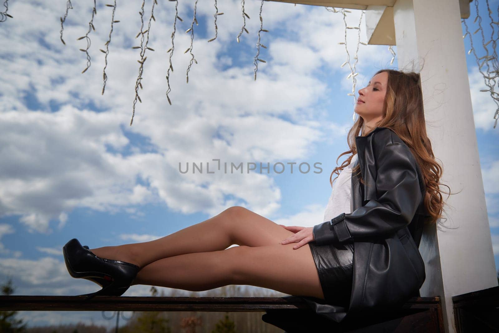 A woman sits gracefully on a ledge dressed in fashionable clothing with a serene expression against a cloudy sky.