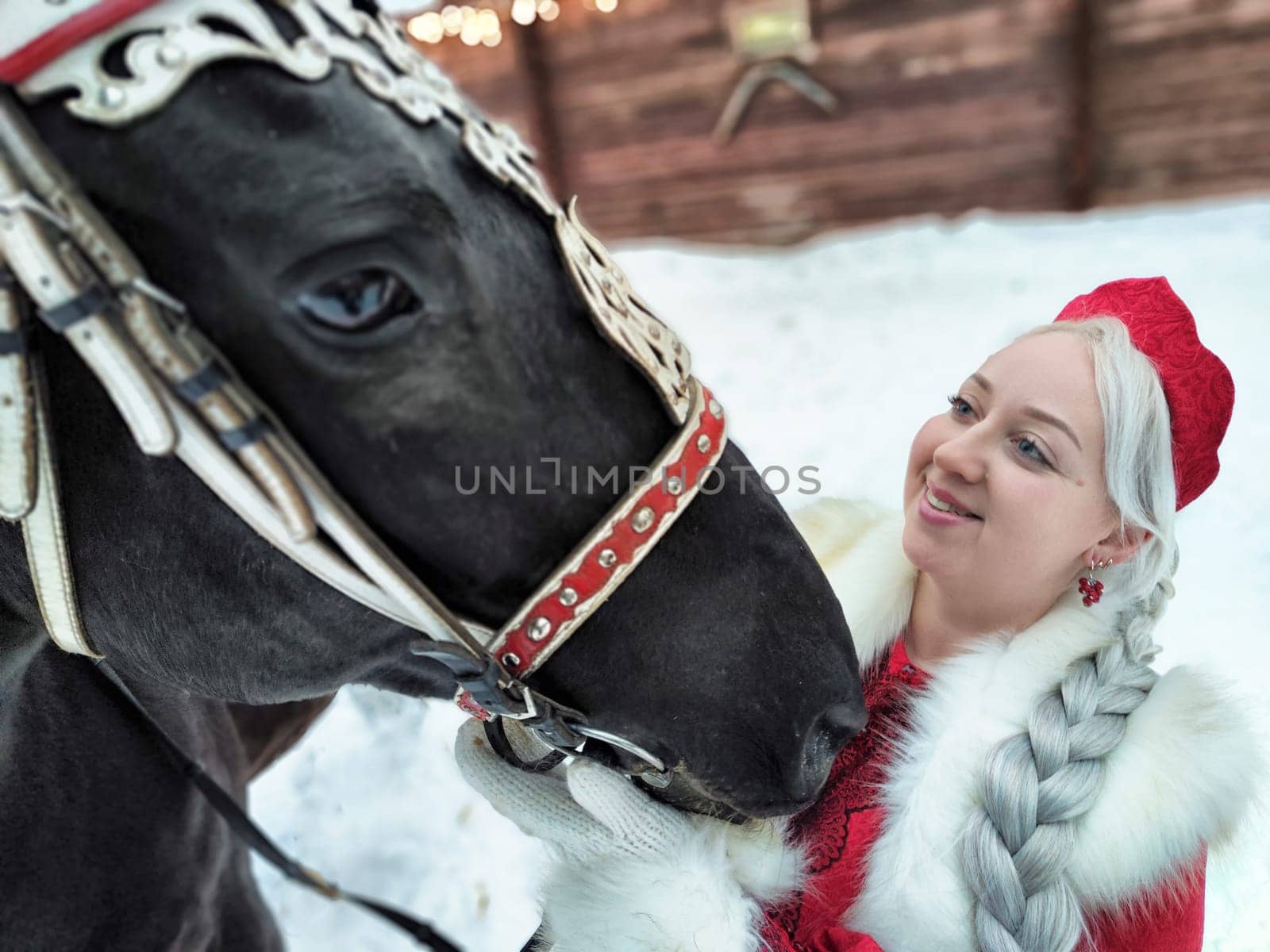 A joyful girl in traditional Russian attire shares a moment with a horse during the Maslenitsa holiday in winter.