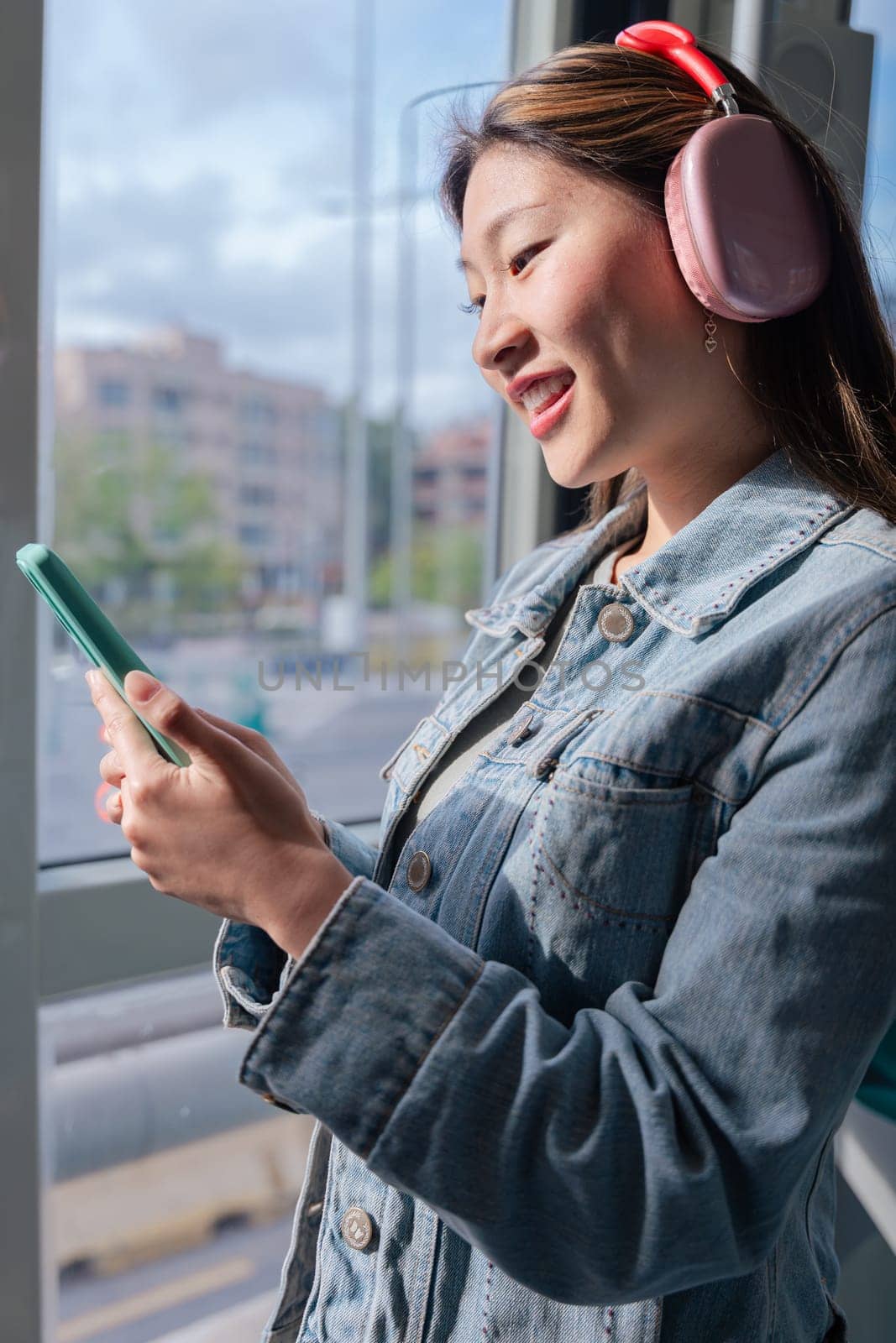 Young chinese woman wearing headphones and denim jacket holding smartphone while looking out the window of a city tram.Peaceful moment during commute, listening to music or thinking calmly.Vertical