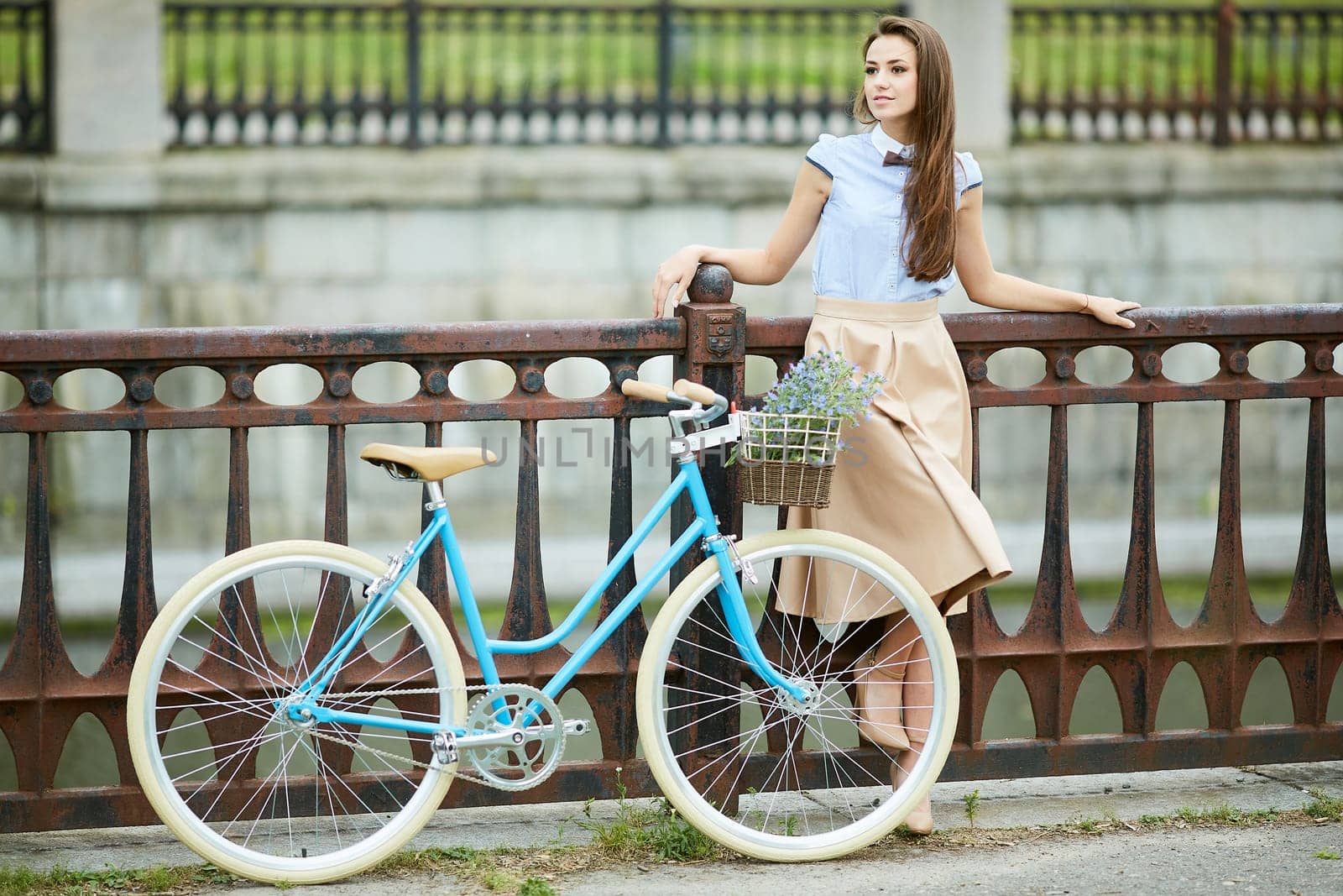 Stylish young woman with long hair smiling, posing with a vintage blue bicycle on a city bridge, wearing an elegant beige skirt and heels
