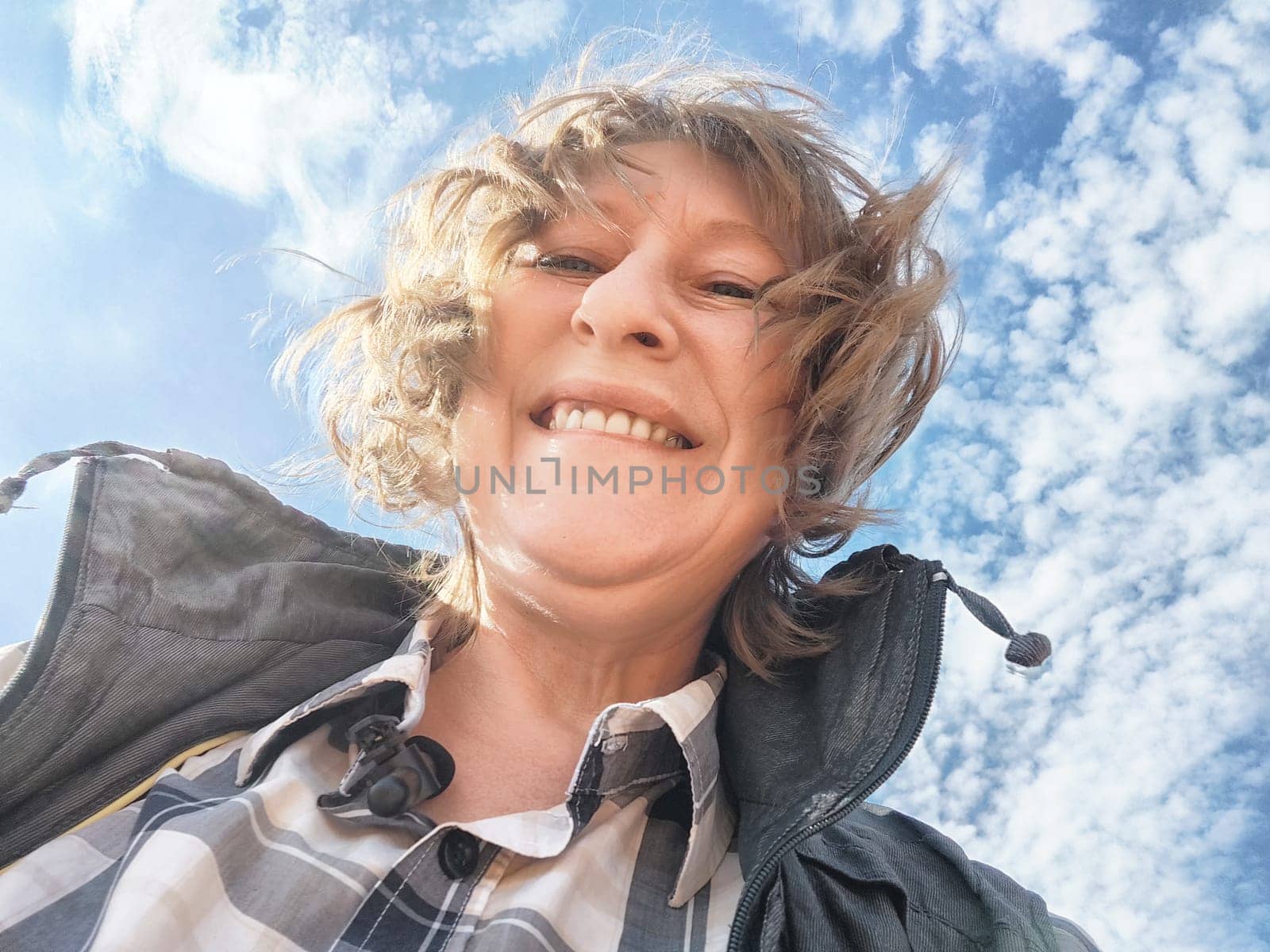 A middle-aged woman shares a joyful selfie under a bright blue sky with fluffy clouds while traveling.