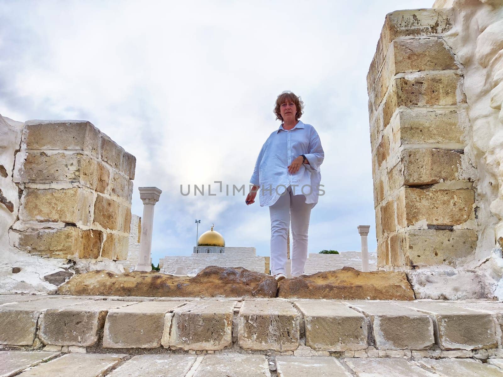 Blonde woman on a sightseeing excursion walks through ancient stone structures, enjoying a travel adventure under cloudy skies.