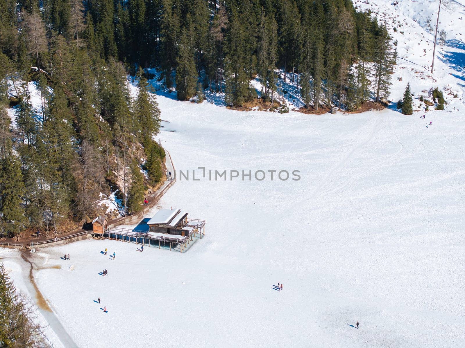 Frozen Braies Lake in the Dolomites, Italy, surrounded by evergreen trees. A wooden boathouse with a walkway sits on the ice, with people nearby.