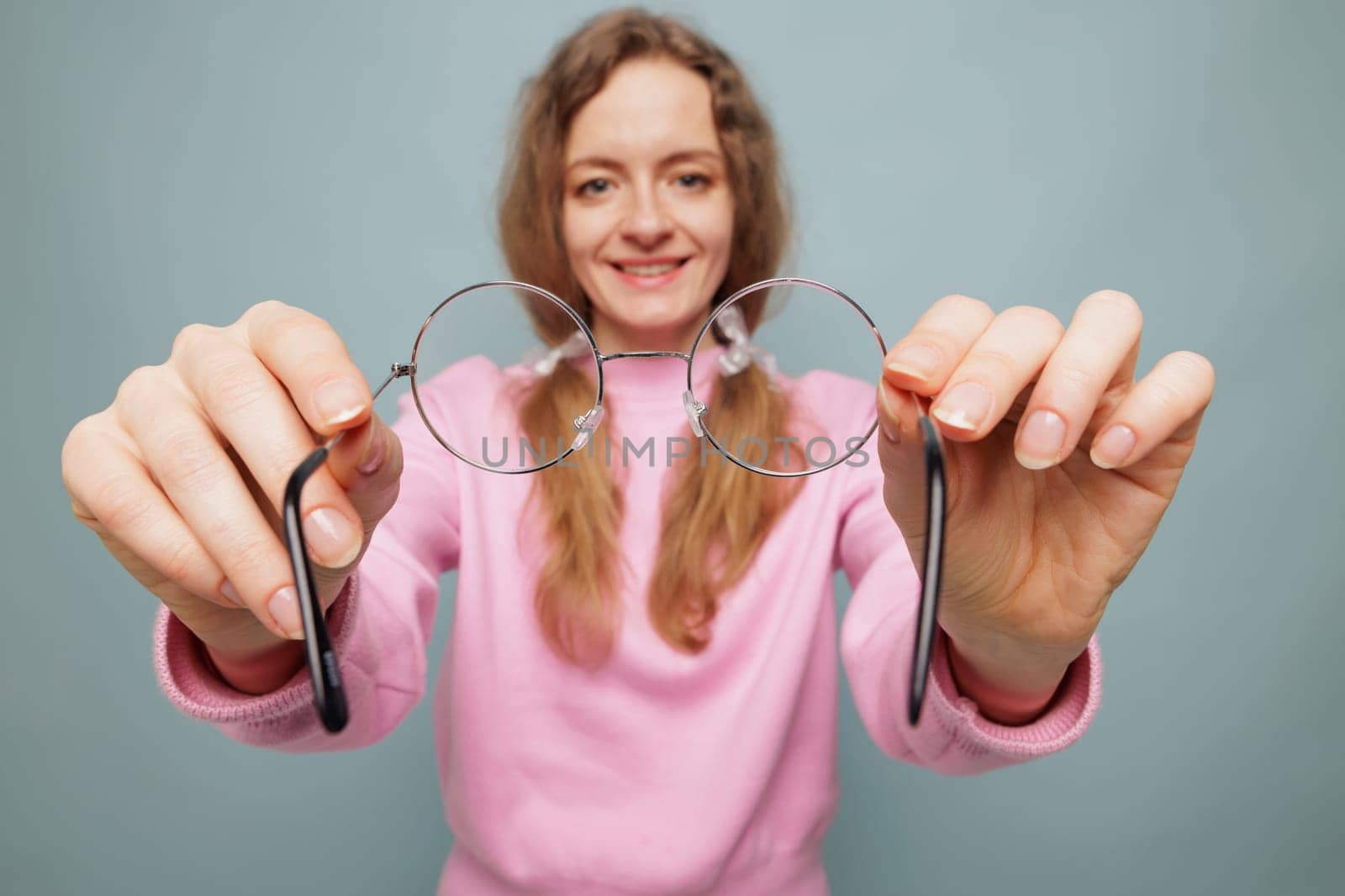A smiling woman in a pink sweater holds a pair of round glasses towards the camera, set against a light blue background.