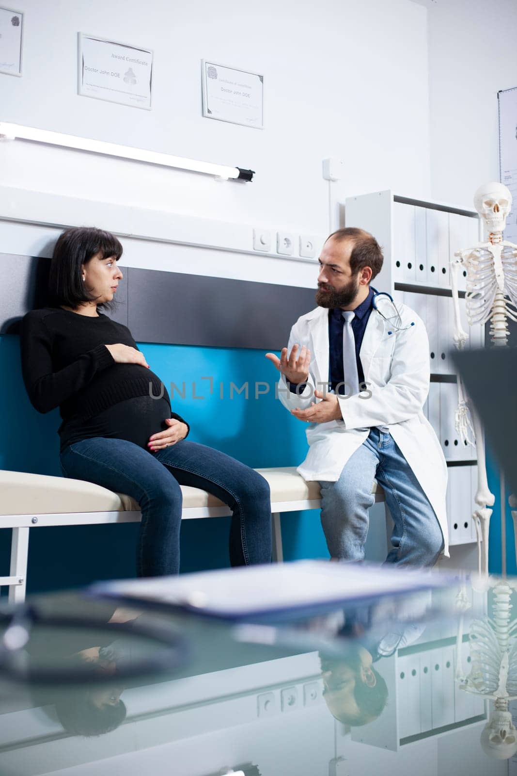 Expectant mother with visible belly bump consults with physician, seated on hospital bed, receiving advice on maternal health. Male doctor explaining prenatal care treatment to pregnant lady in clinic