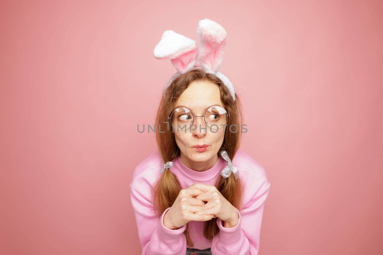 Playful woman in pink sweater and bunny ears making a funny expression. Quirky Easter concept, humorous portrait, festive mood, and spring vibes