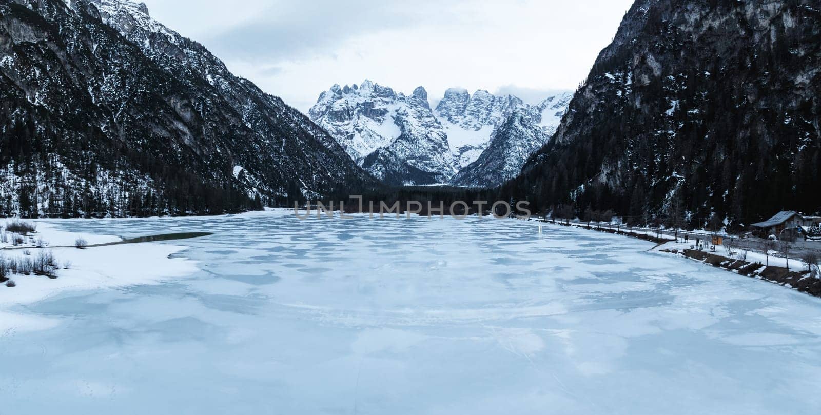 A frozen lake with natural ice patterns, surrounded by snow covered alpine peaks and dense forests. A small structure is visible near the shoreline.