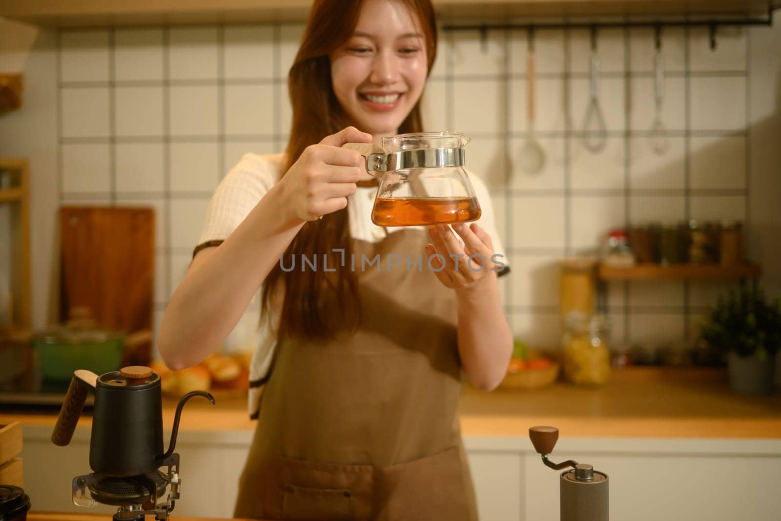 Smiling woman holding a glass carafe filled with freshly brewed coffee, standing in a cozy kitchen.