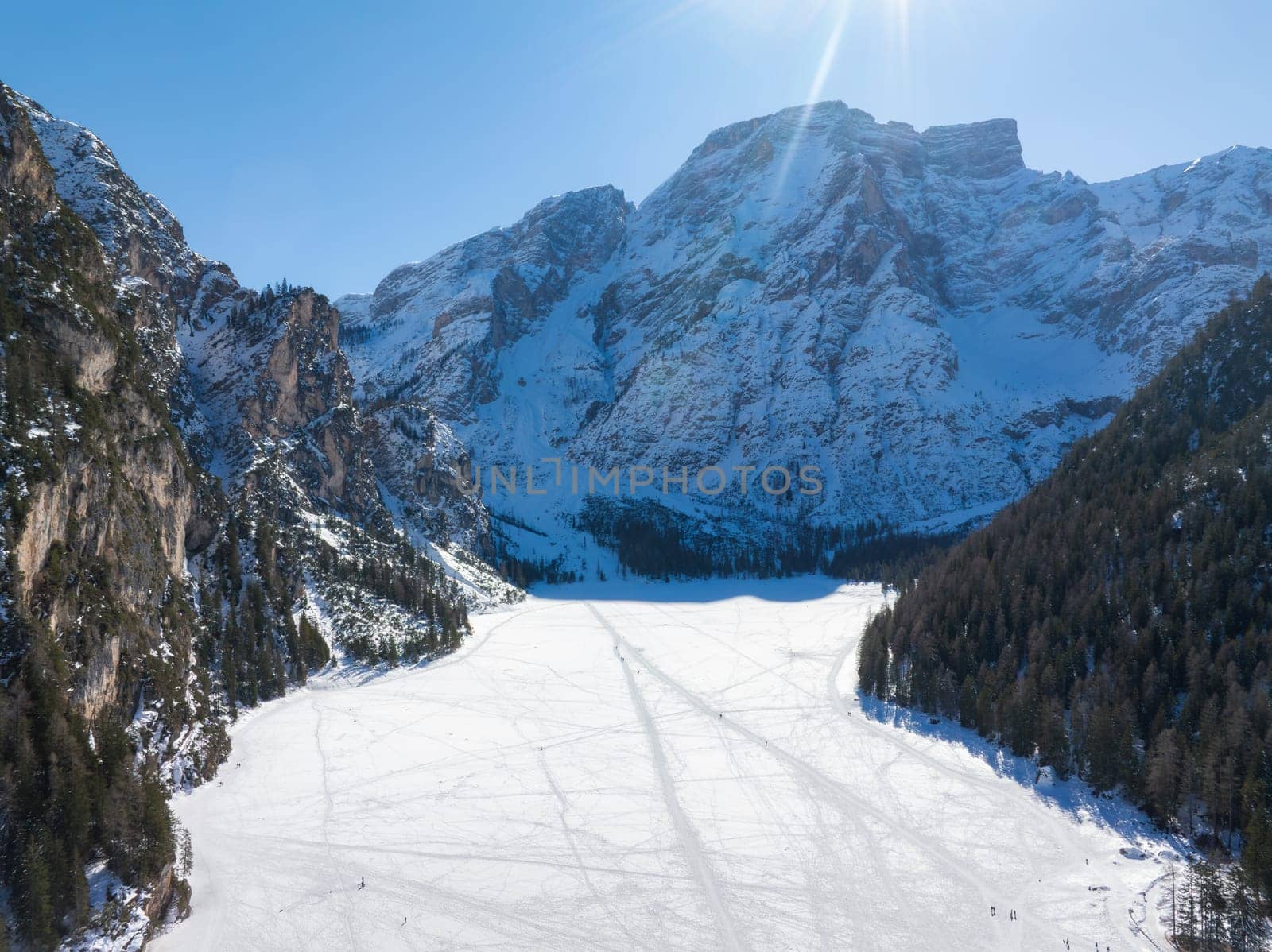 Aerial view of Braies Lake in winter, frozen and snow covered with tracks on its surface. Snowy mountains and evergreen forests surround the lake.