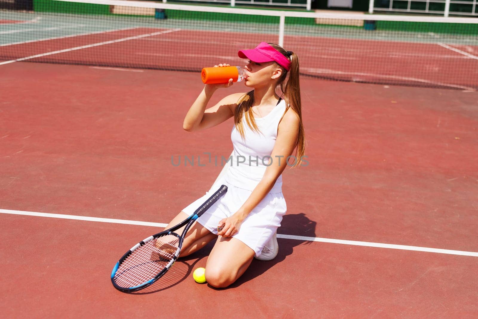 Young female tennis player drinking water, holding racket and ball, wearing sportswear and visor, rehydrating after practice on an outdoor court, tennis game