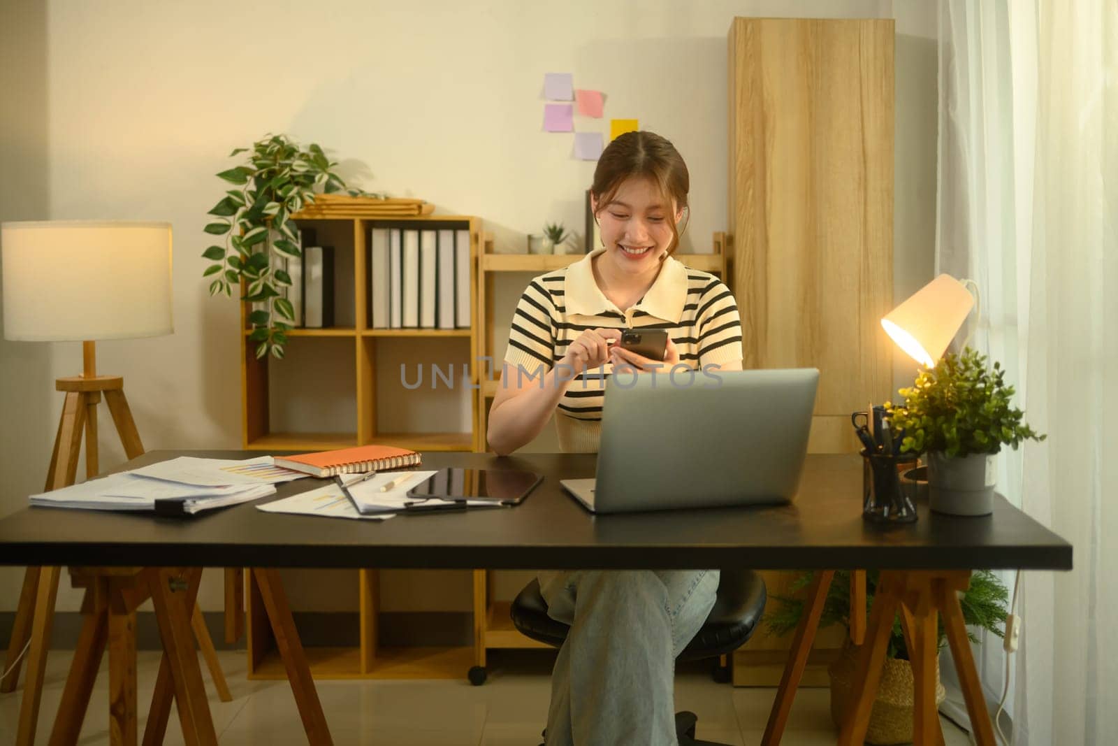 Smiling young woman using smartphone while working at home office desk with laptop.