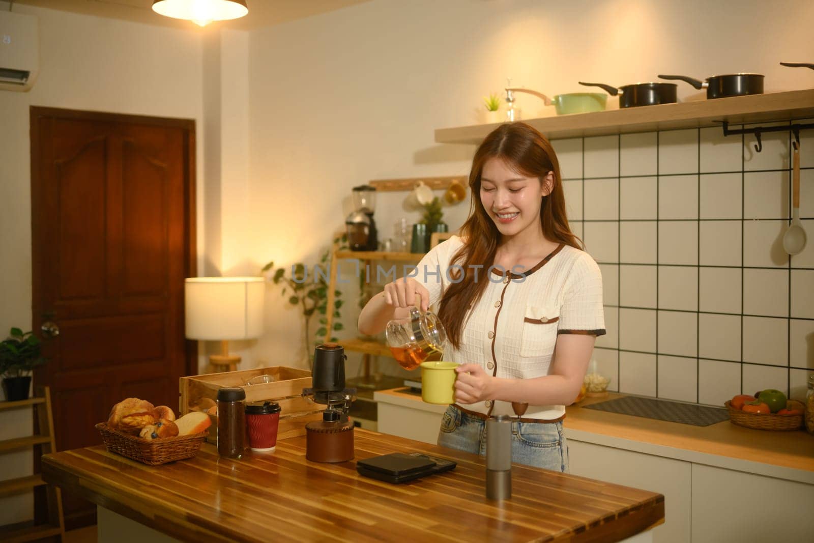 Happy asian woman pouring freshly brewed coffee into a mug in her warm kitchen.