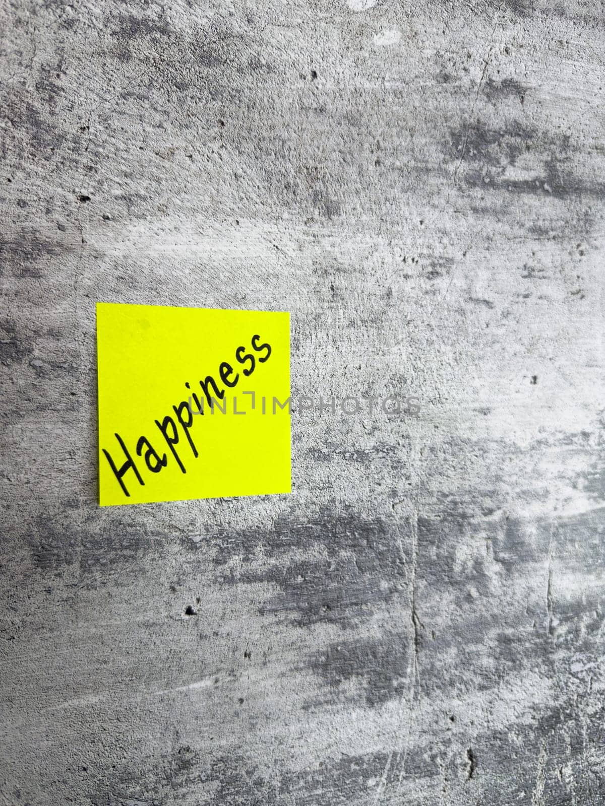 Bright yellow sticky note inscribed with the word Happiness, displayed on a textured grey background, representing the essence of joy and the pursuit of a fulfilling, positive life